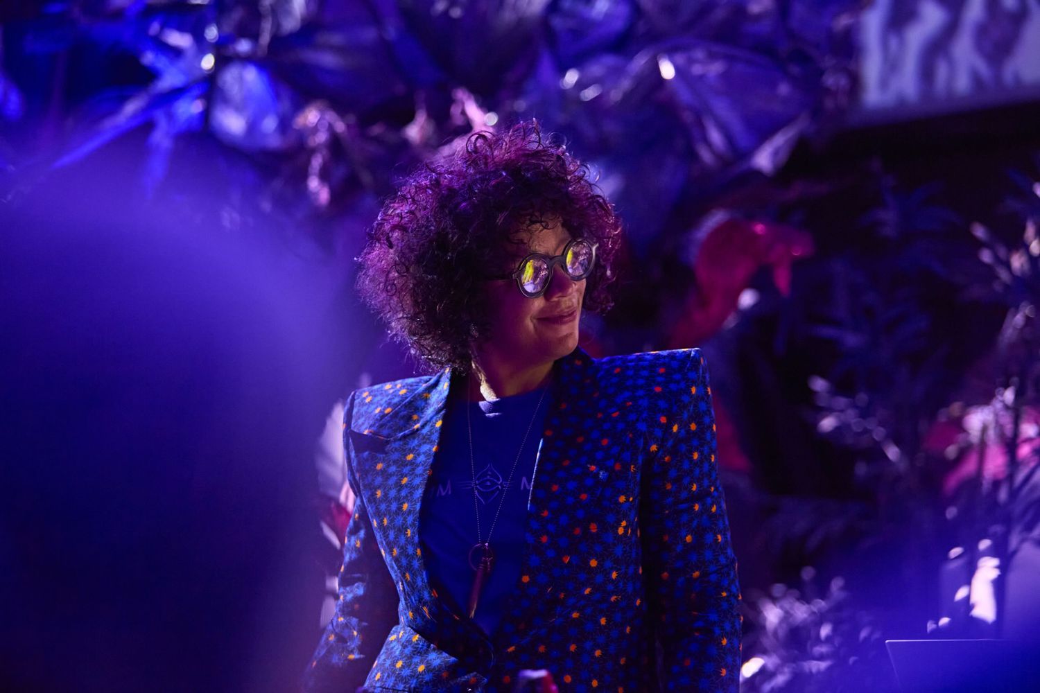 Person with curly hair wearing blue sequined blazer and glasses in colorful purple lit setting.