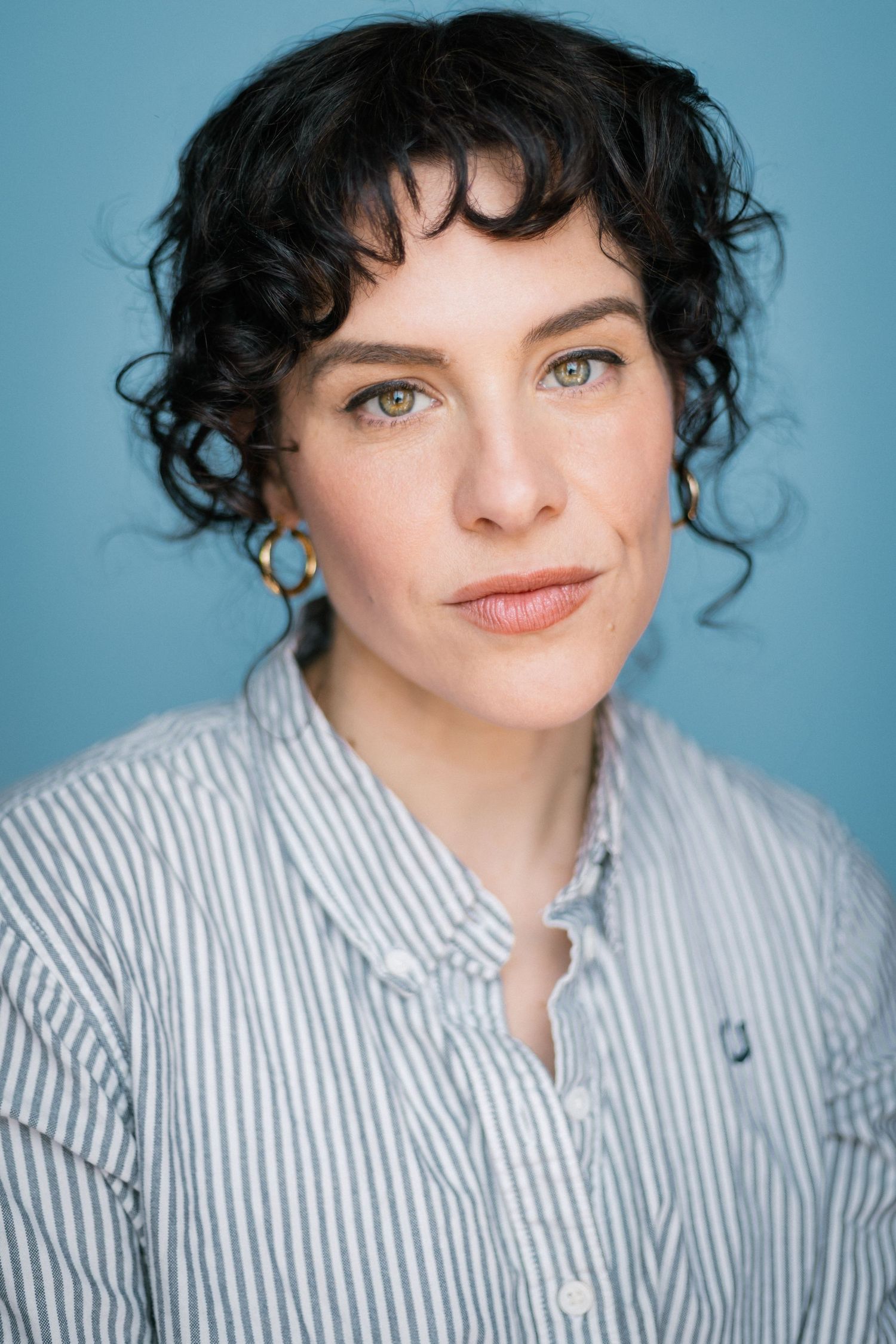 Woman with short curly black hair and green eyes wearing a striped shirt on blue background.