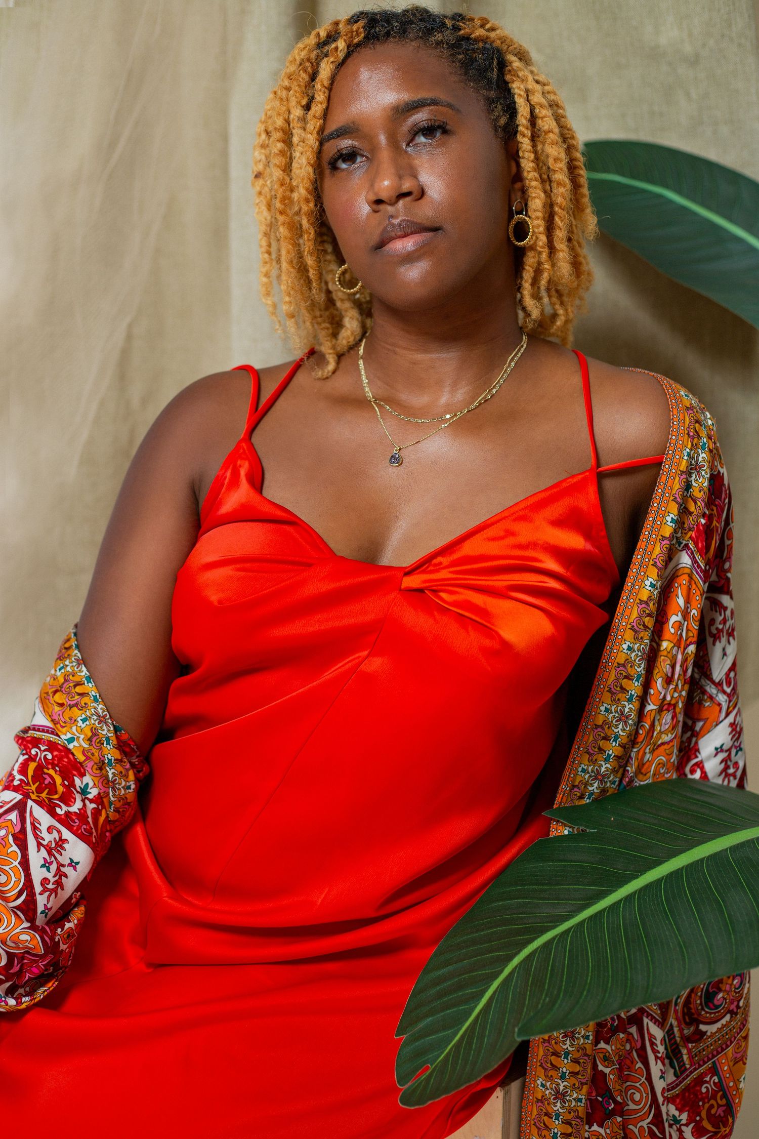 Woman in red satin slip dress with patterned kimono posing with tropical leaves.