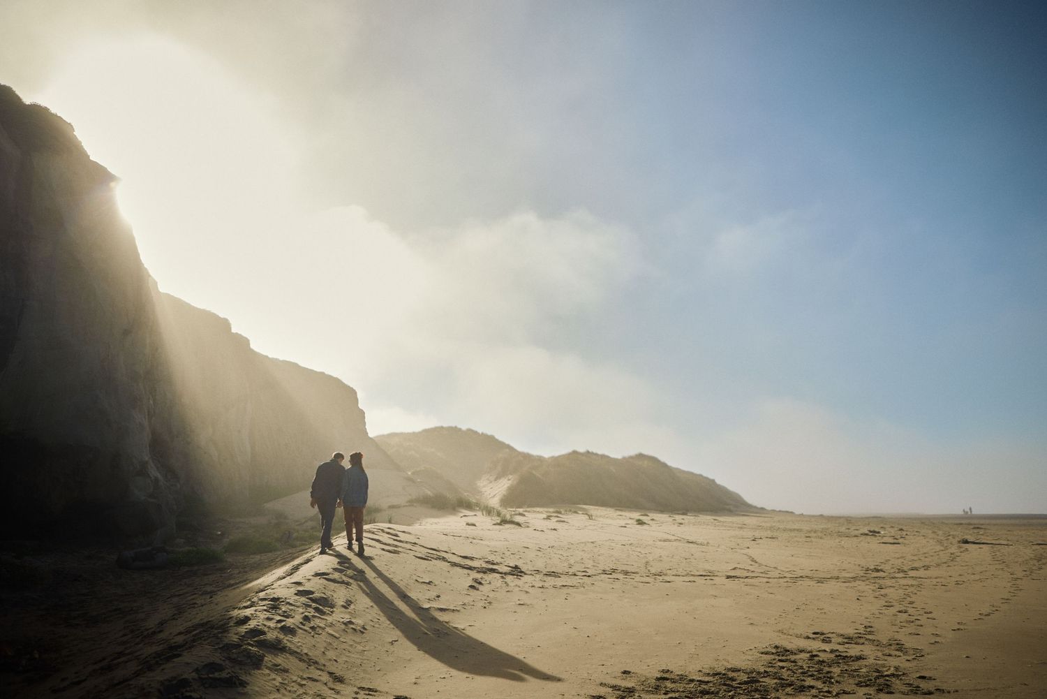 A couple walks hand in hand along a sandy beach with dramatic cliffs and misty sunlight.