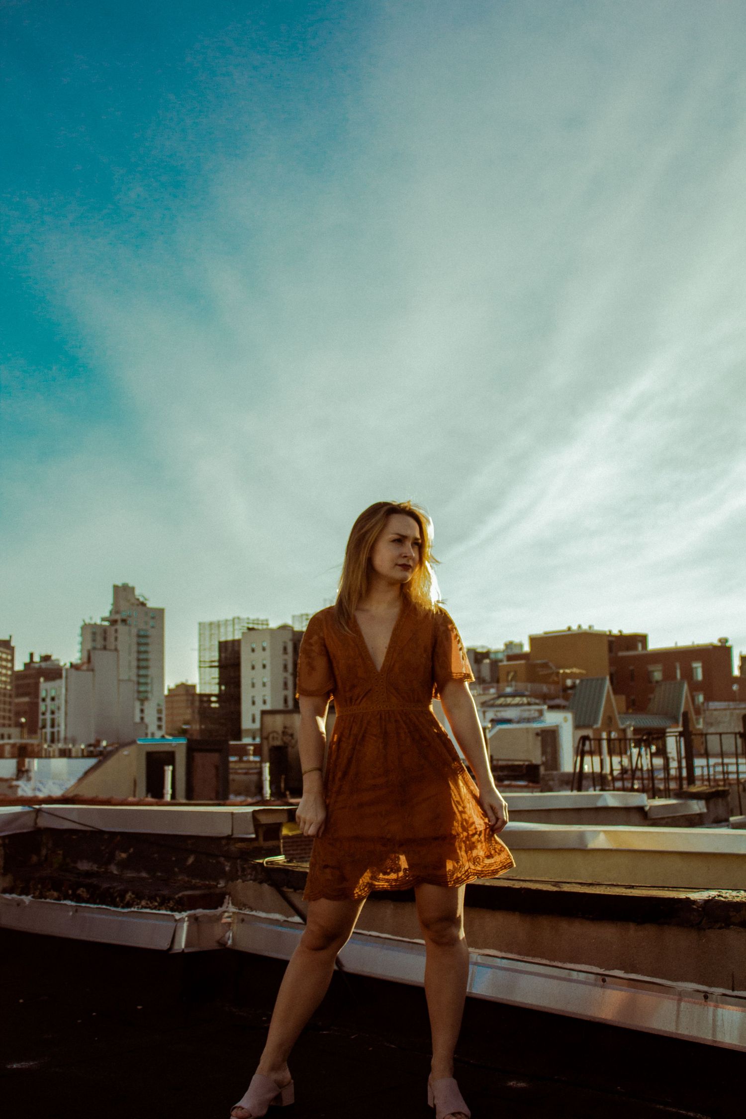 Blonde woman in rust-colored wrap dress stands on rooftop with city skyline at golden hour.