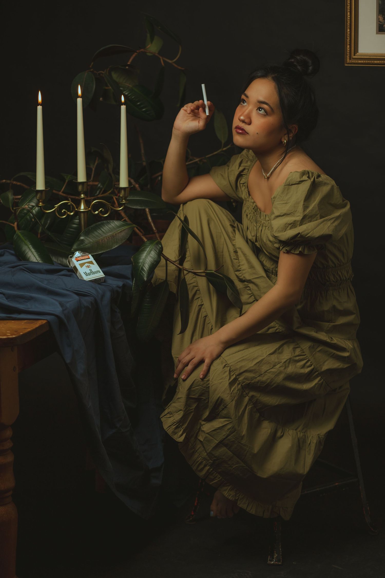 Woman in olive green dress sitting beside candles and plants in a dark, moody studio setting.