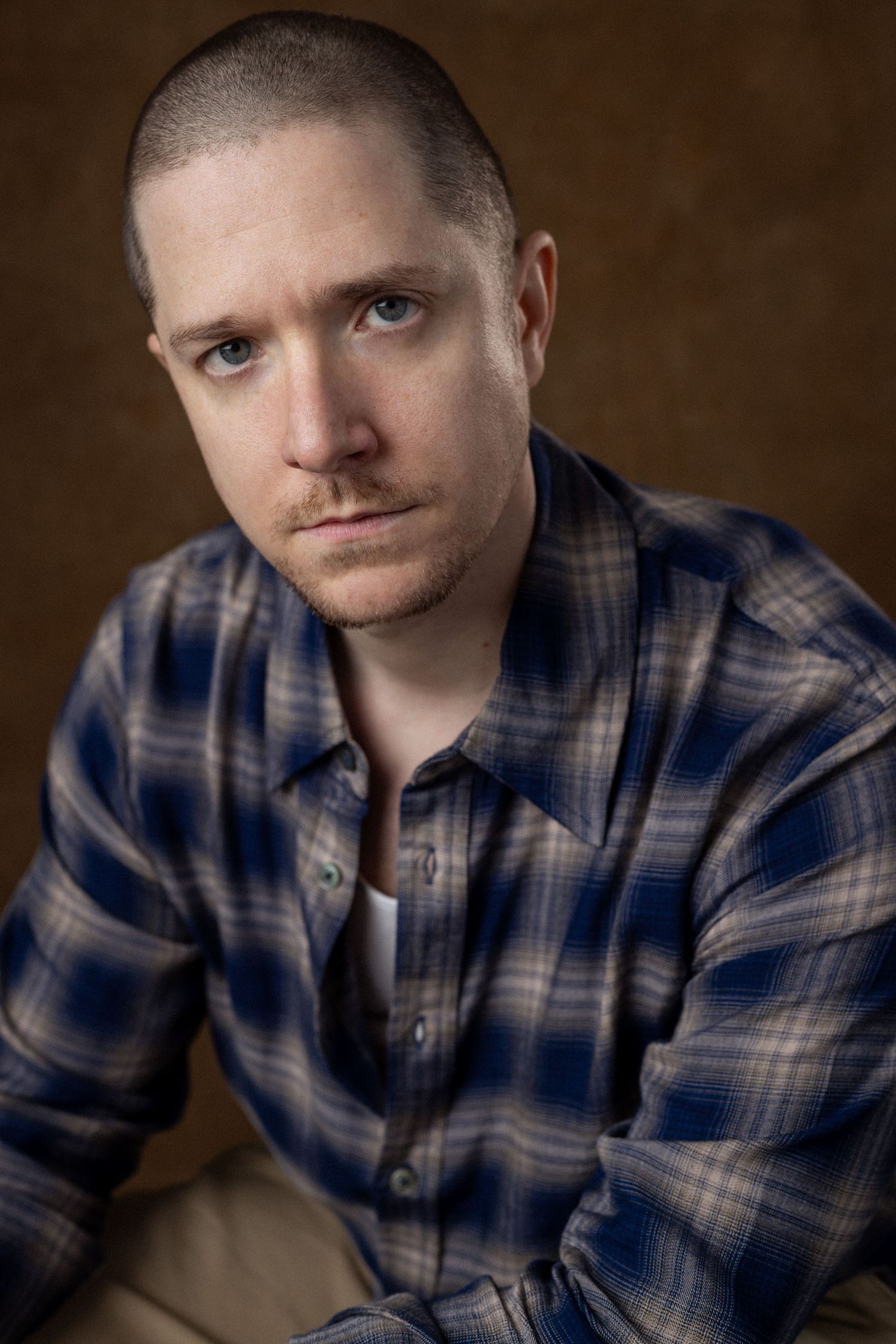 Young man with buzz cut wearing blue plaid flannel shirt, posing for portrait against brown background.