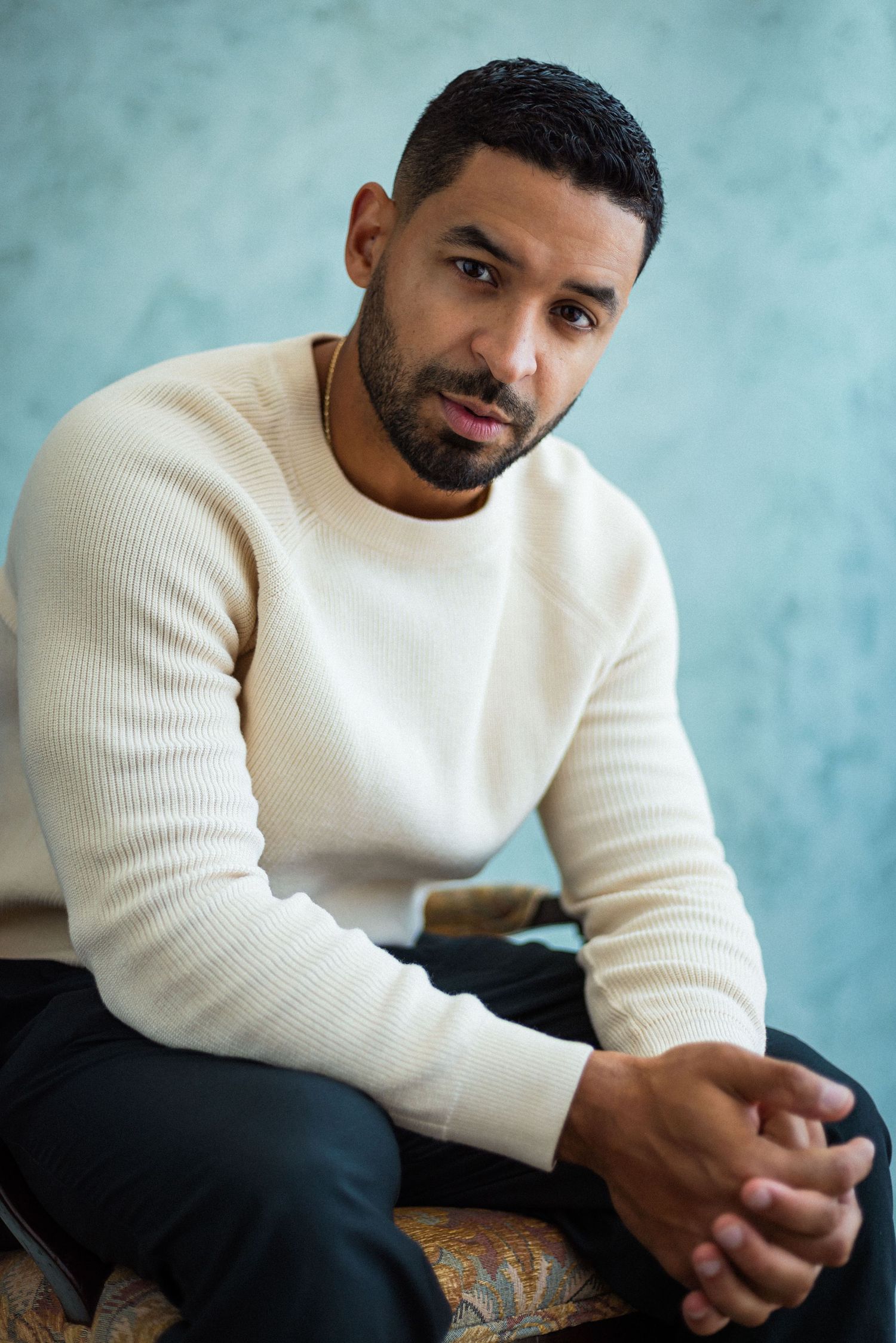 A young man with a beard wearing a cream knit sweater sits with clasped hands against a blue textured wall.
