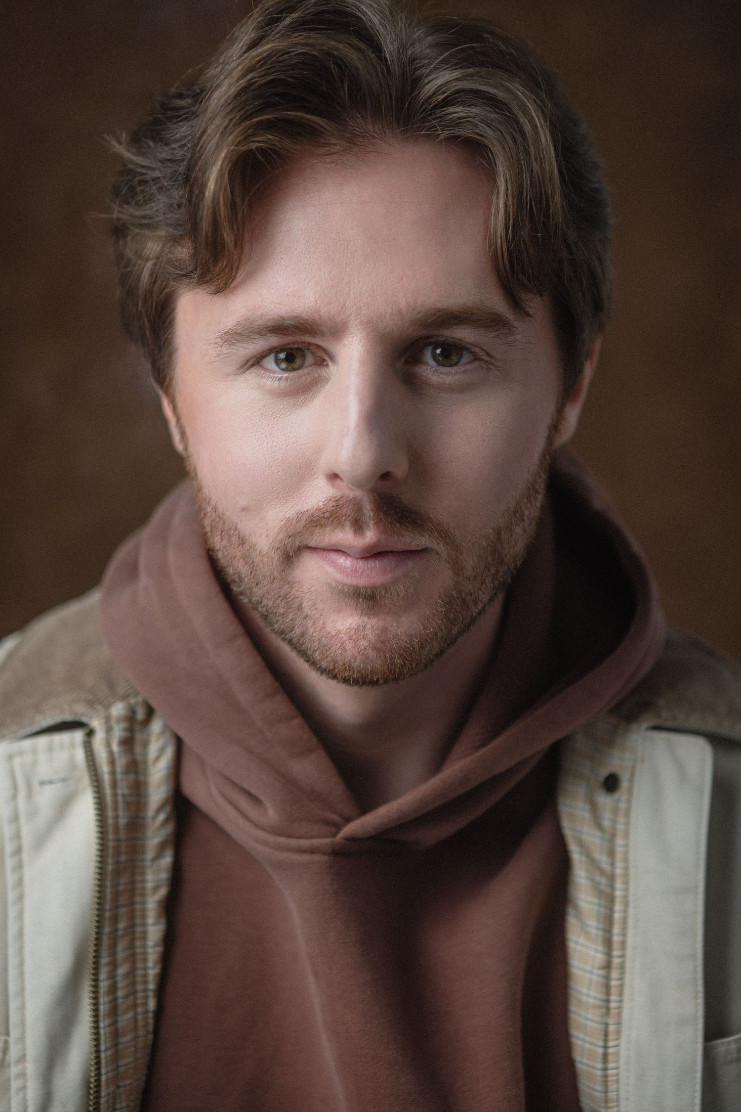 Close-up portrait of a young man with light brown hair, stubble beard, wearing a brown hoodie and beige jacket.