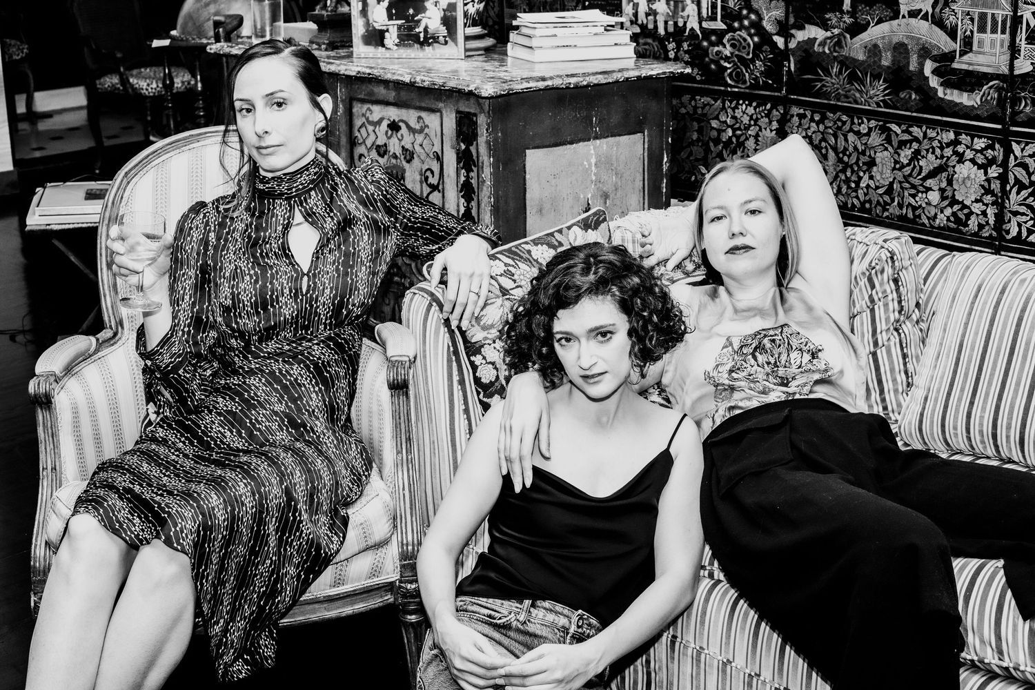 Three women pose together on a vintage sofa in a black and white artistic portrait photograph.
