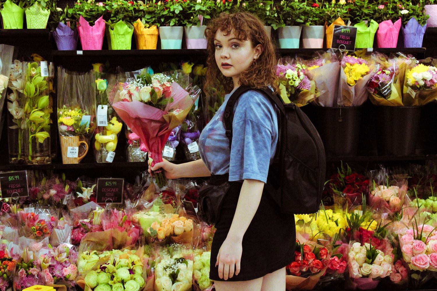 Young woman with curly red hair holds flowers while standing in a colorful flower shop.