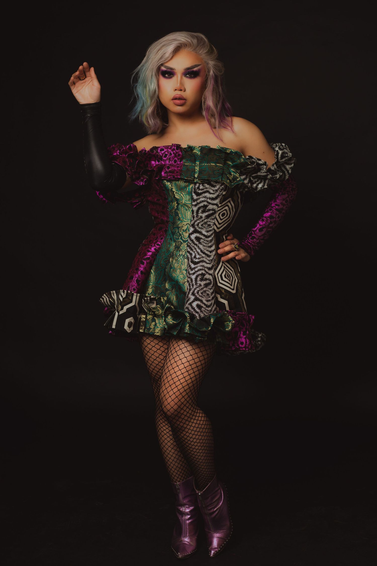 Drag performer in colorful sequined off-shoulder mini dress with fishnet stockings and purple boots poses dramatically.
