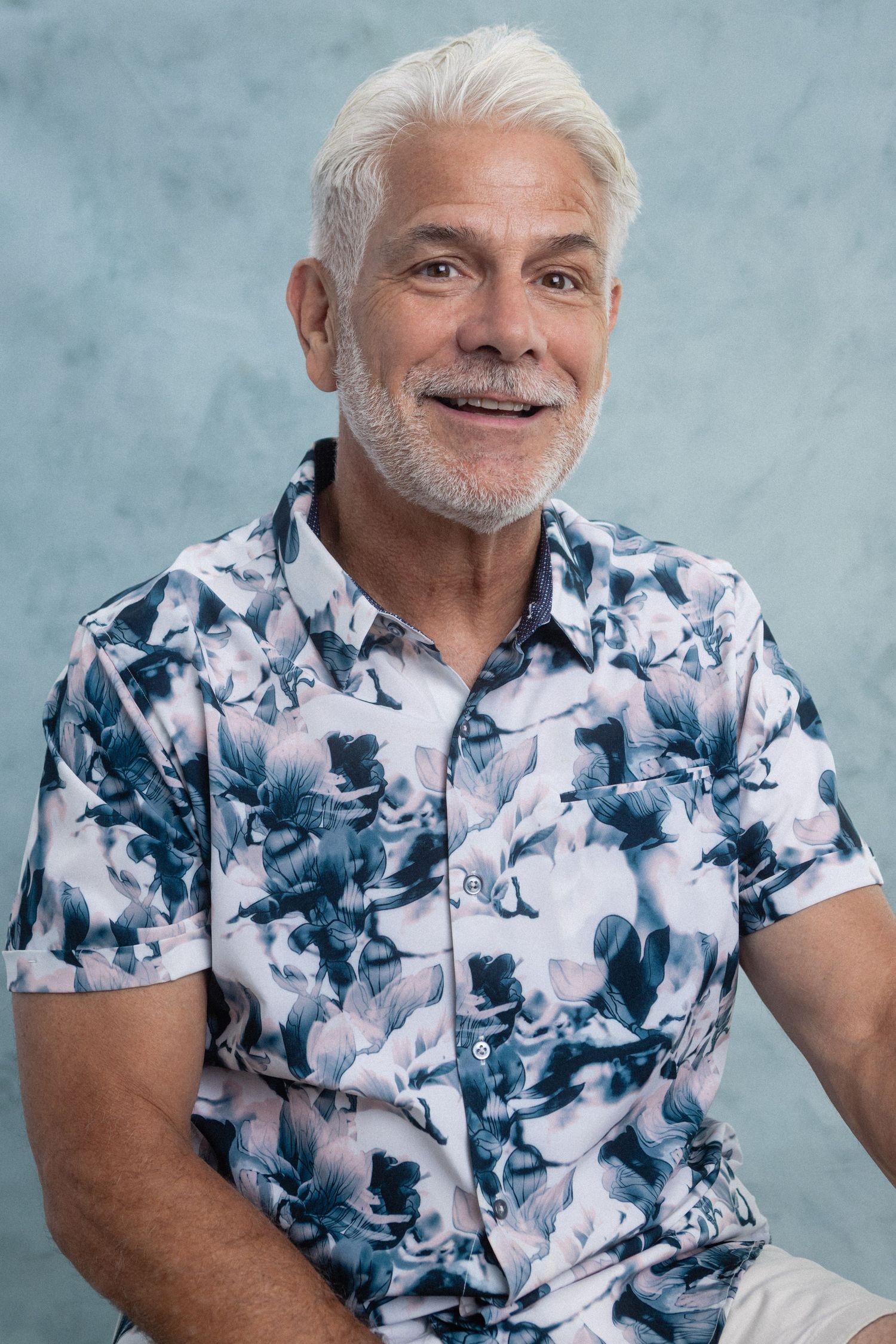 Smiling older man with white hair and beard wearing a floral shirt, posed against a blue background.