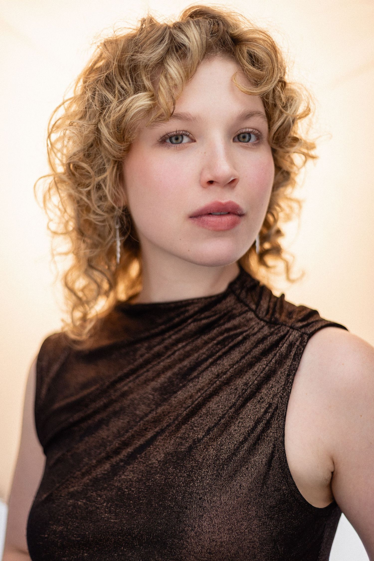 Young woman with curly blonde hair wearing a dark sleeveless top, posing against a warm beige background.