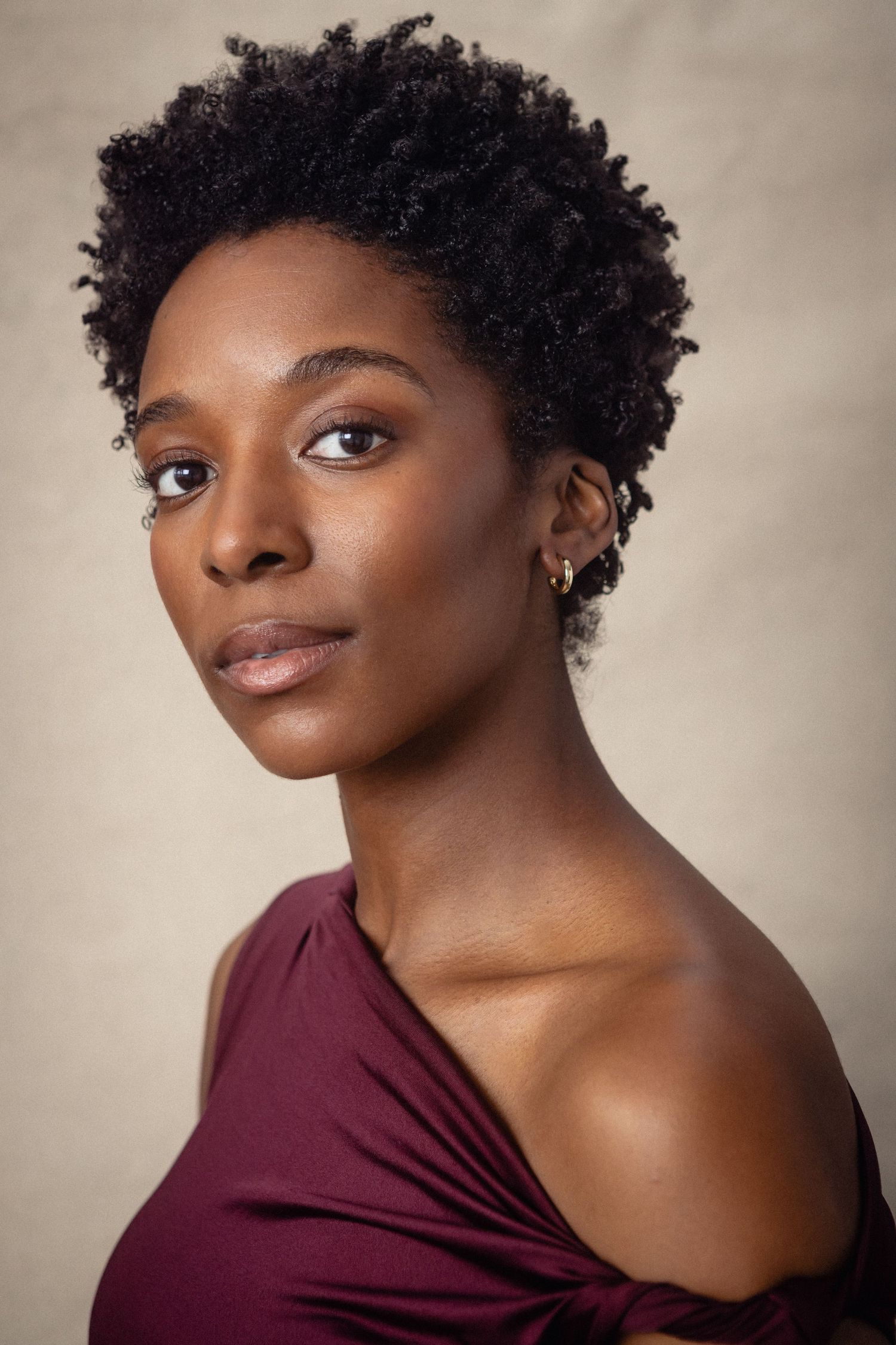 A young Black woman with natural short curly hair wearing a burgundy off-shoulder top poses against a neutral background.