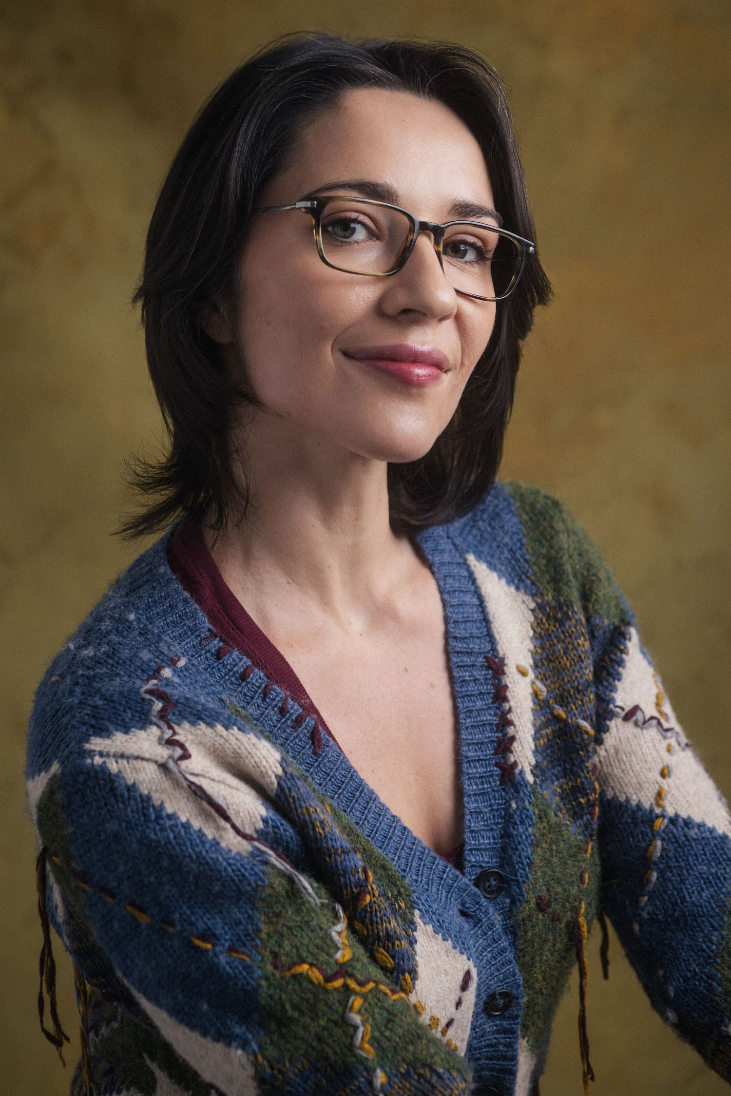 Woman with dark hair and glasses wearing a colorful patterned cardigan sweater, smiling against a golden background.