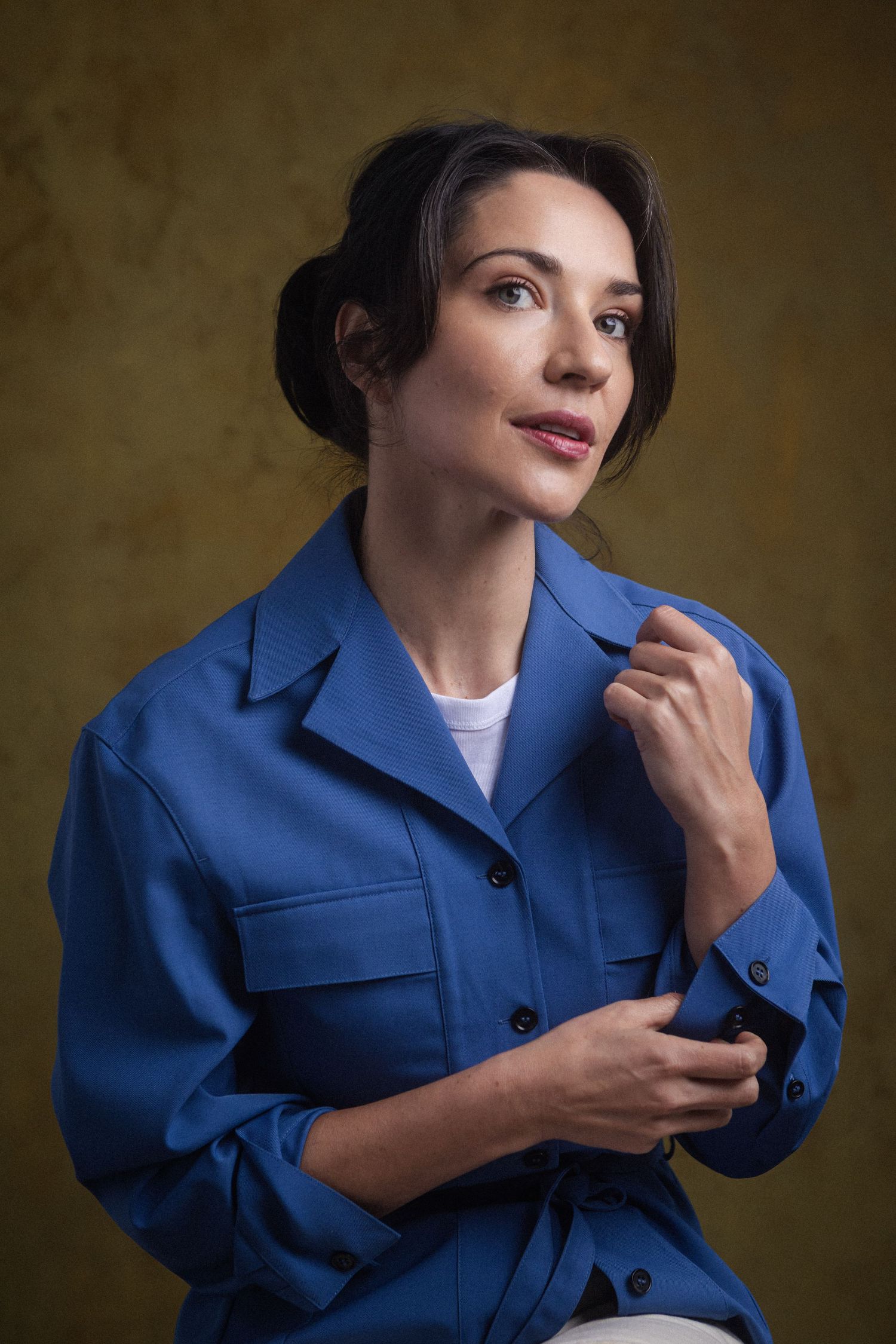 A woman in a blue jacket with dark hair in a bun poses thoughtfully against a dark background.