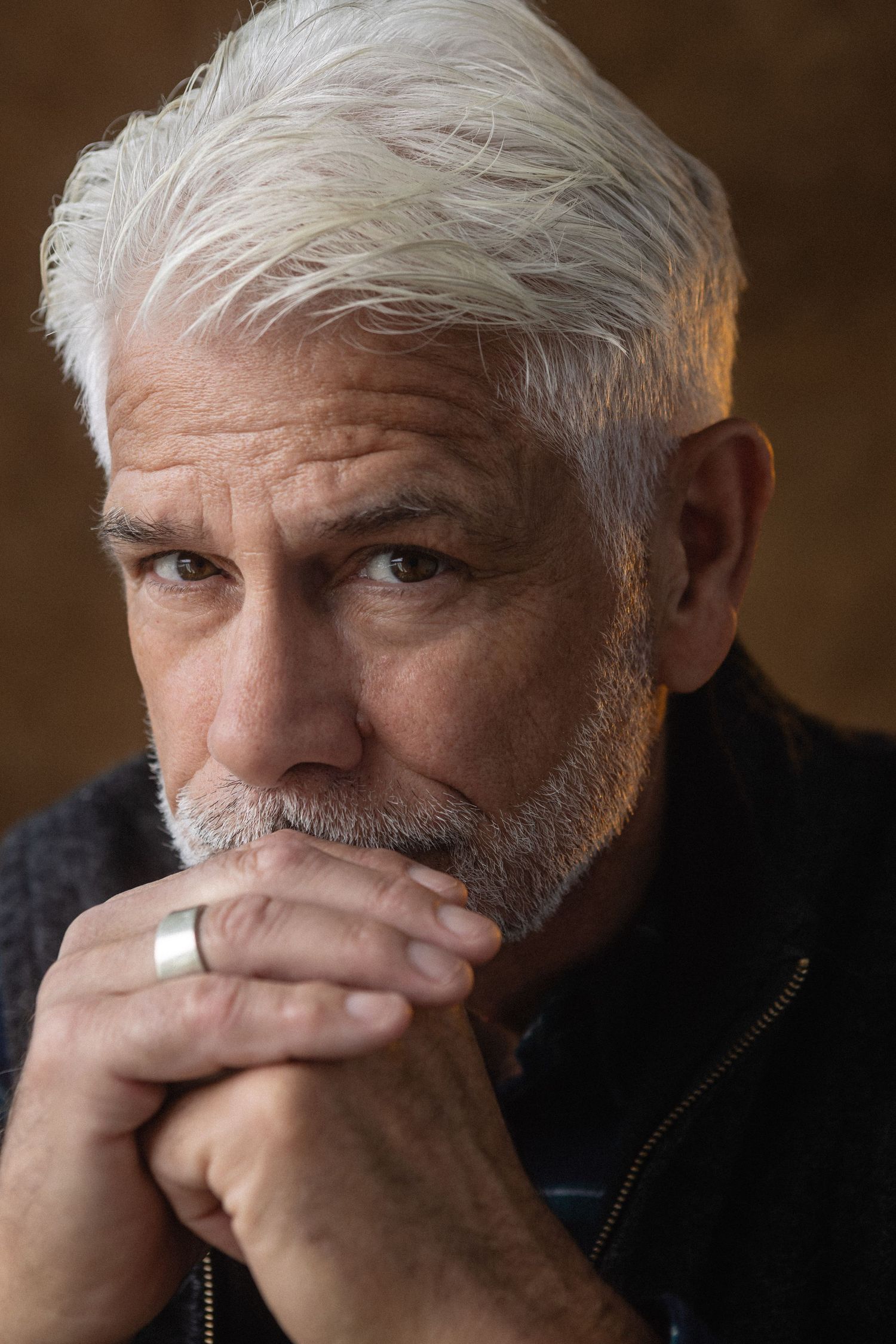 Thoughtful older man with white hair and beard resting chin on hands, wearing a silver ring, against dark background.