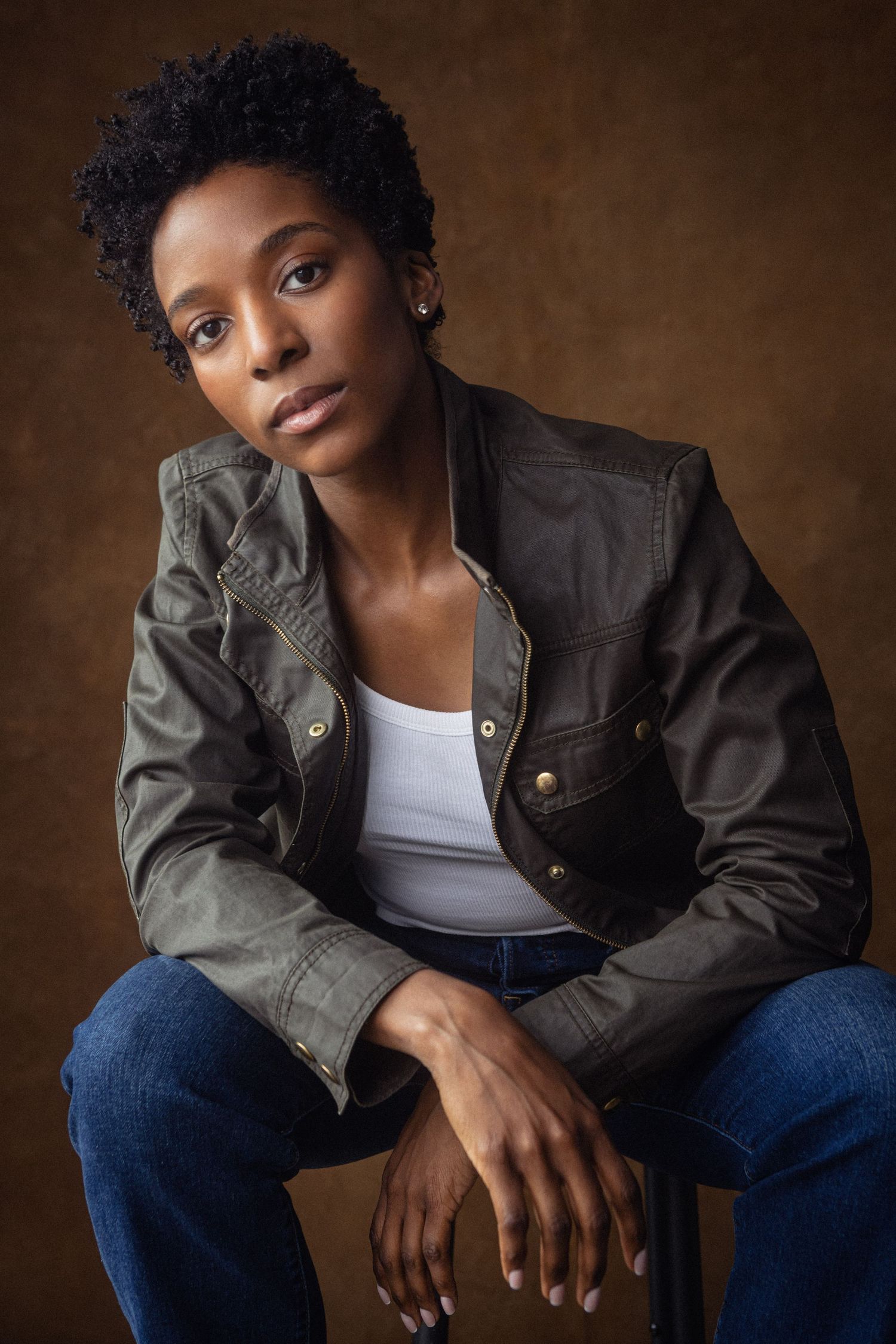 Young Black woman in olive jacket and jeans sits confidently on stool against brown studio background.