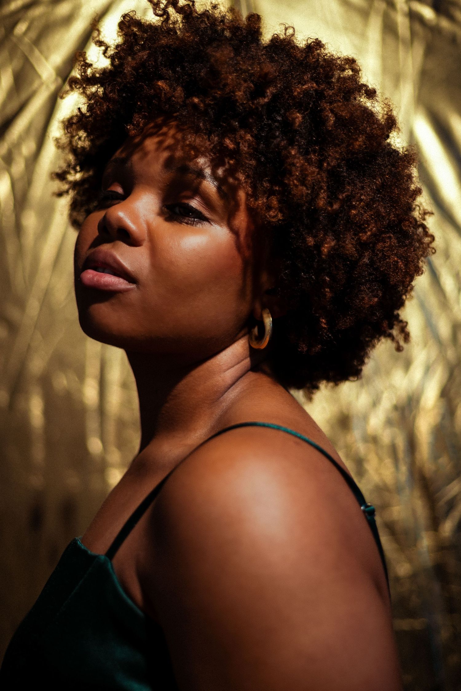 A Black woman with a natural afro hairstyle poses confidently against a golden background wearing a green spaghetti-strap top.