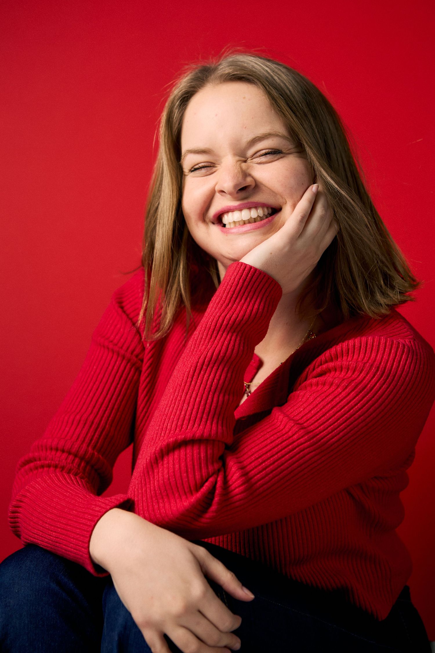 A cheerful young woman in a red ribbed sweater laughing against a vibrant red background.