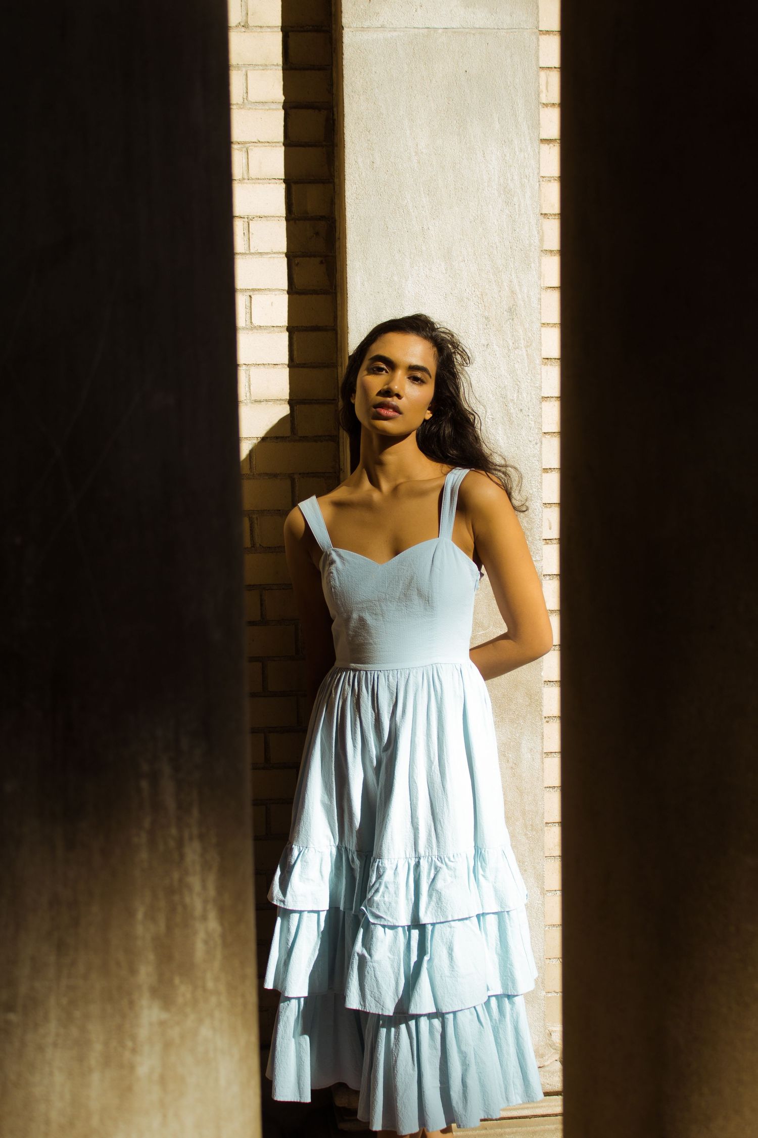 Woman in light blue ruffled sundress stands between shadowed columns in warm sunlight.