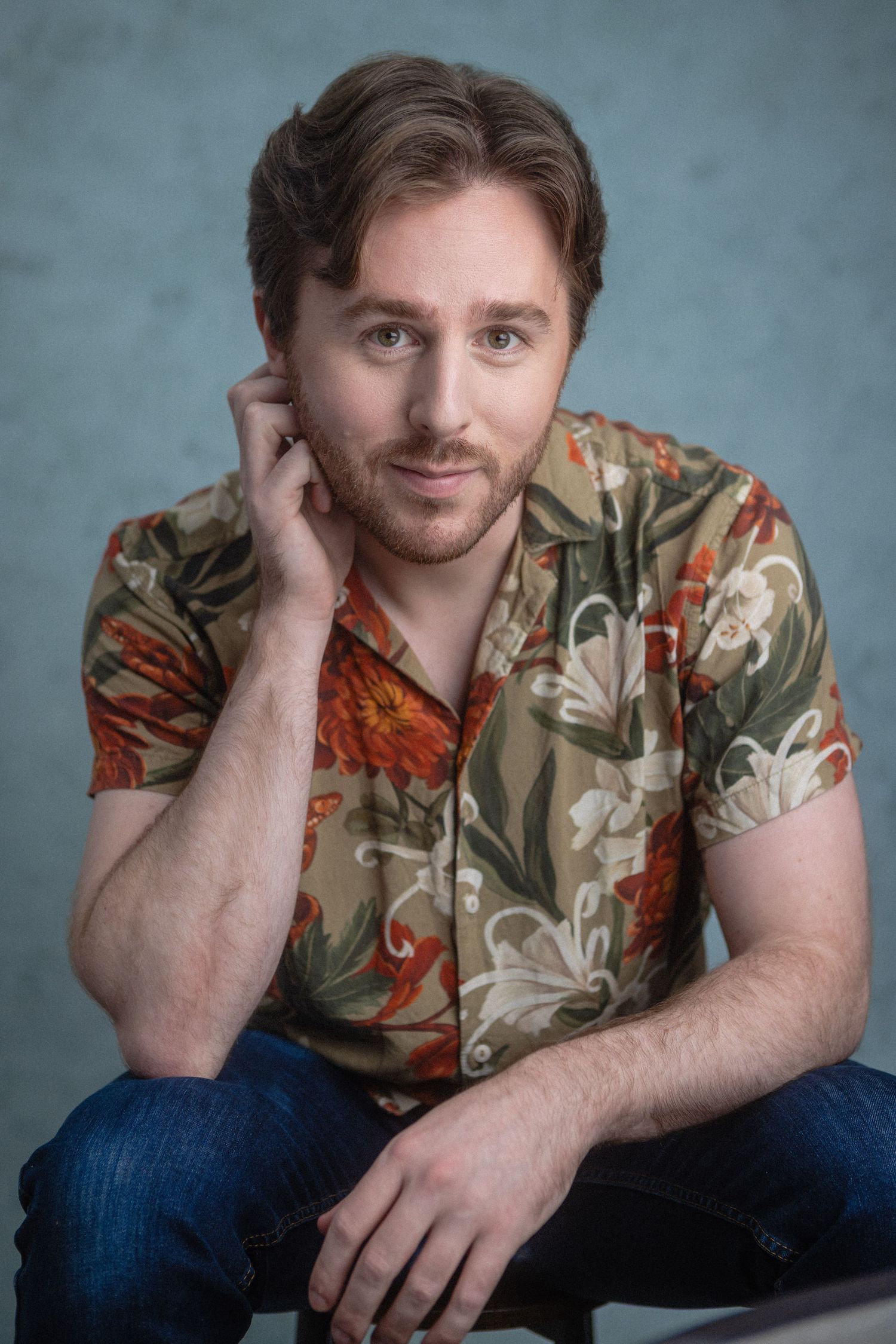 Young man with beard wearing floral Hawaiian shirt sitting relaxed against blue-gray studio background.