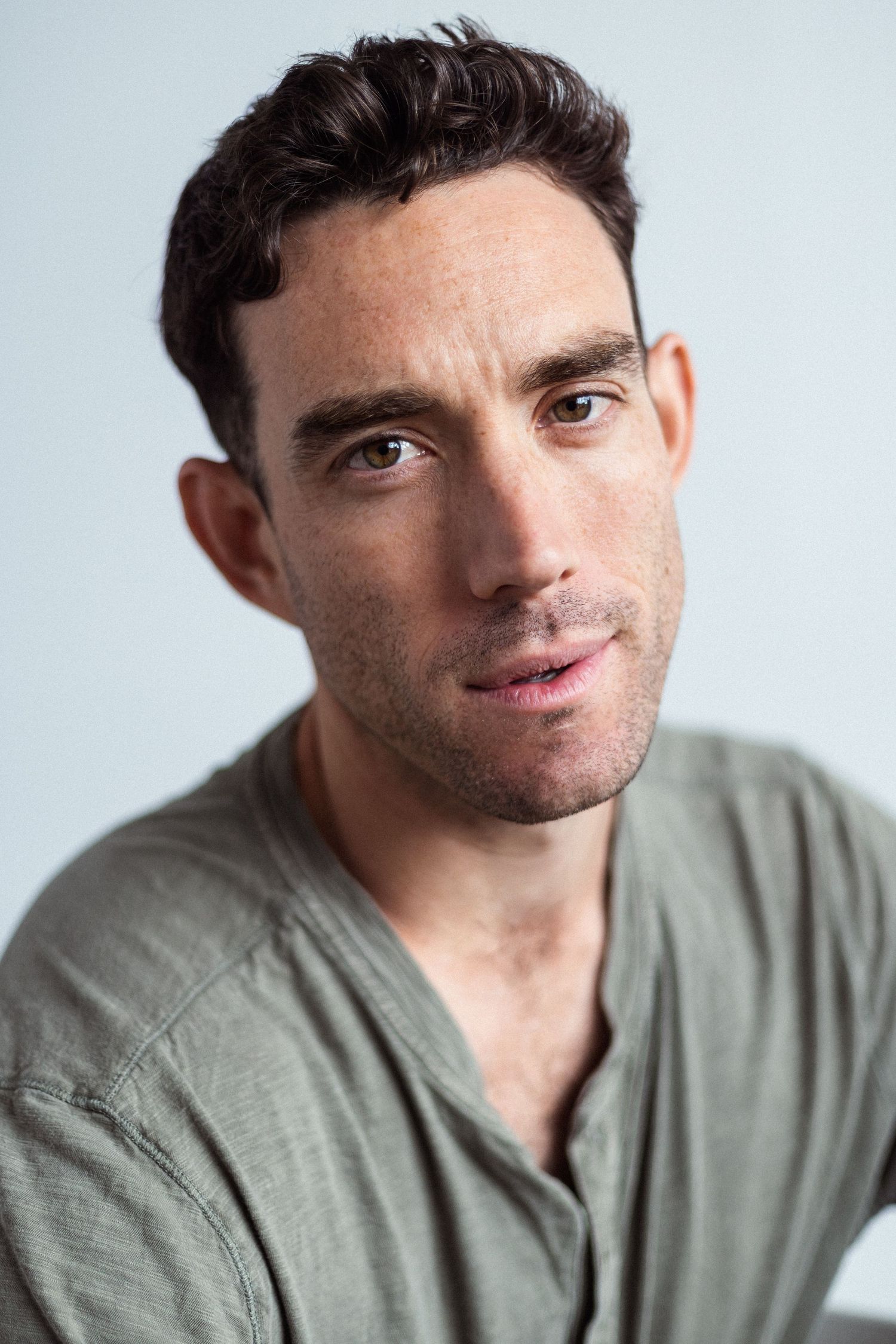Close-up portrait of a young man with dark hair wearing a sage green linen shirt against a light gray background.