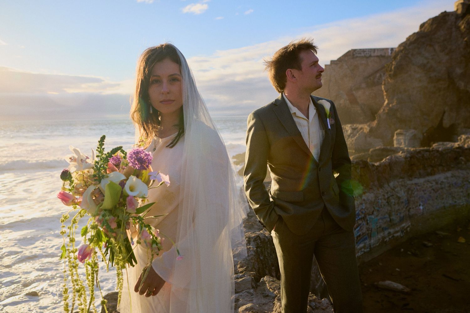 Bride in white veil holding colorful bouquet with groom in gray suit posing on rocky ocean cliffside at sunset.