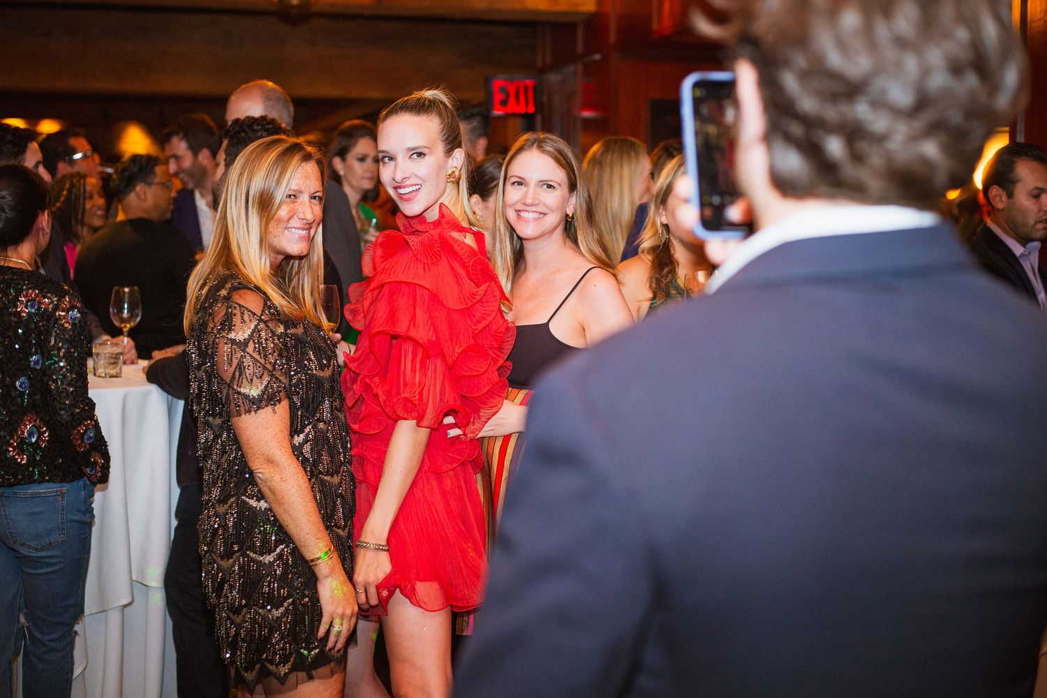 Three women posing for a photo at an indoor social event, one wearing a red ruffled dress.