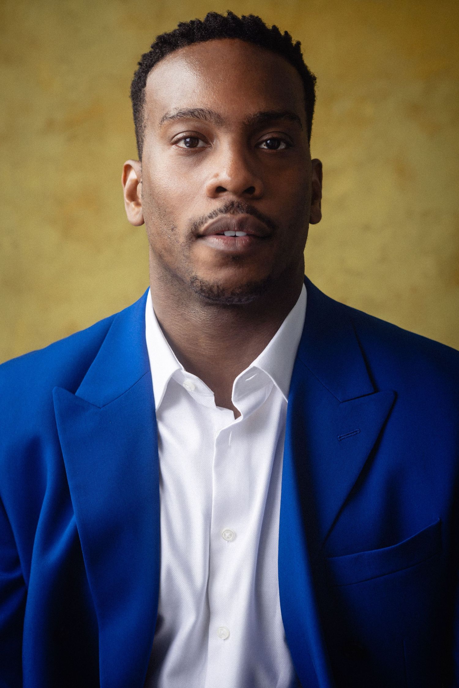 Young Black man in blue blazer and white shirt poses confidently against a yellow background.