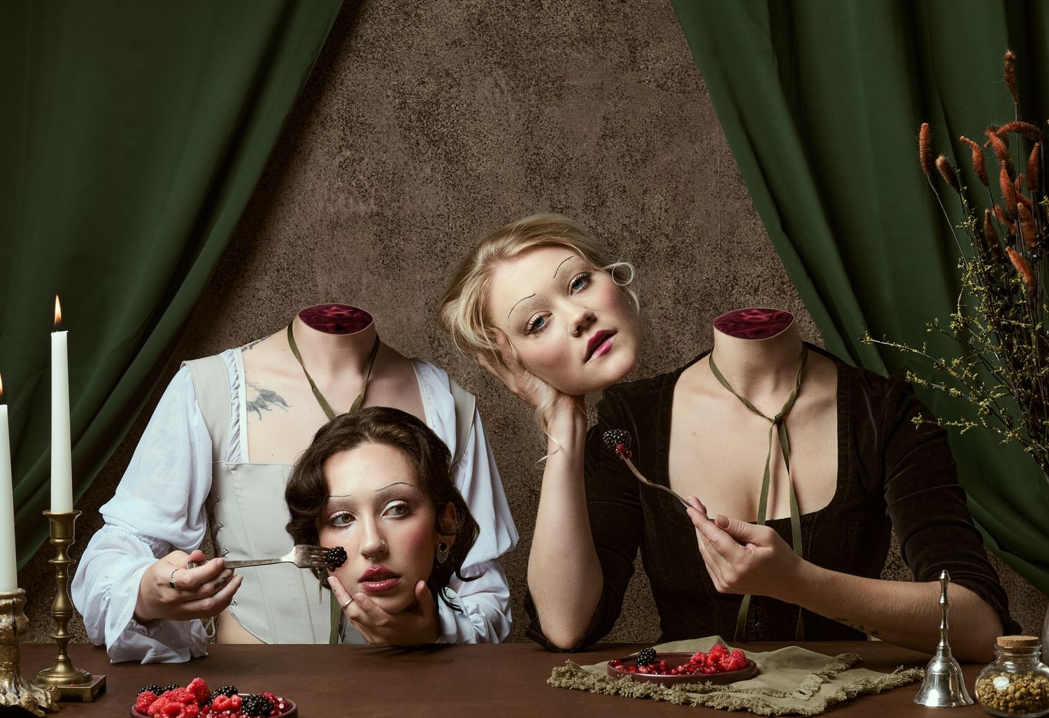 Two women pose dramatically at a table with candles, holding detached heads in a surreal, Renaissance-inspired theatrical scene.