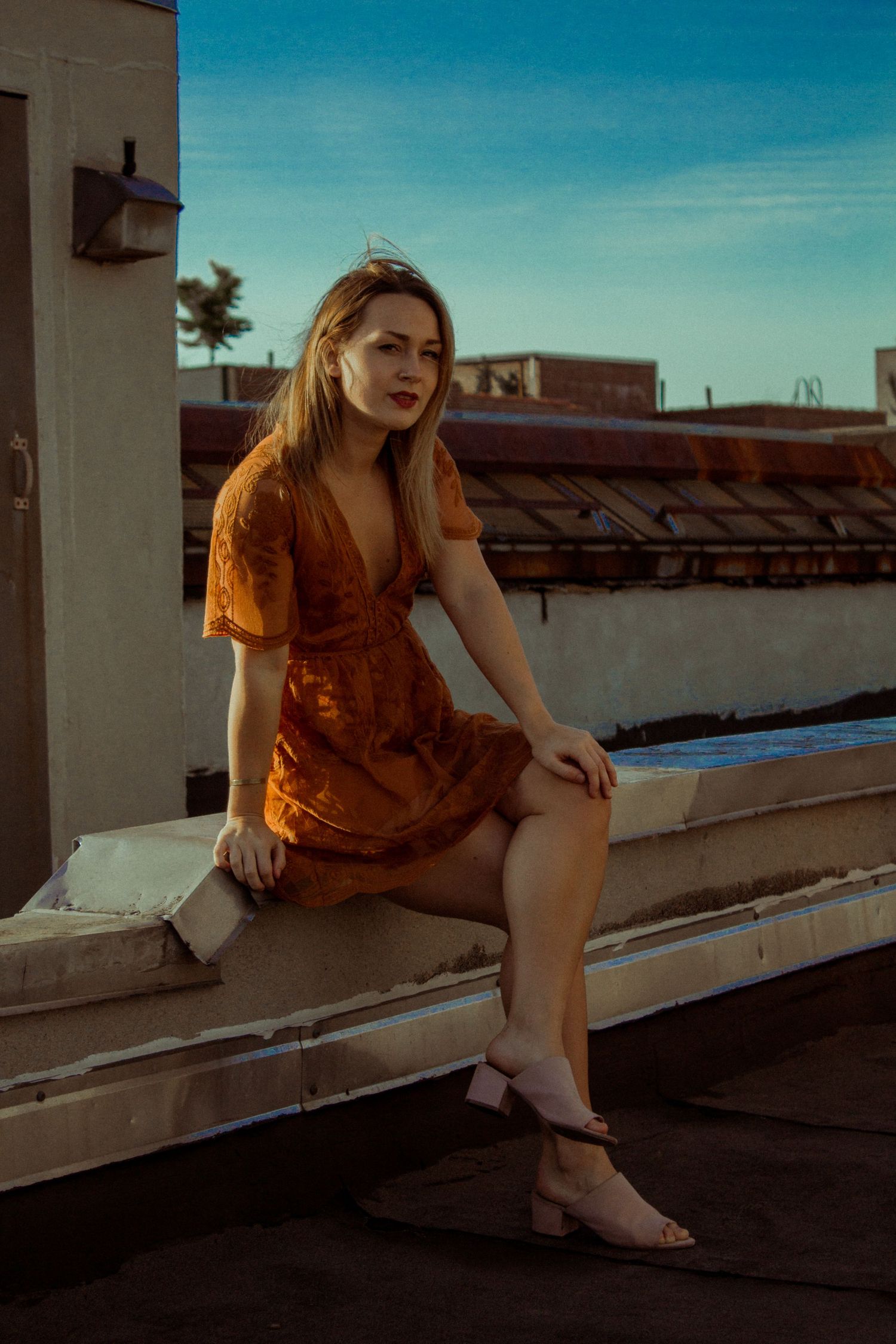 Young woman in orange mini dress sitting on rooftop ledge at golden hour.