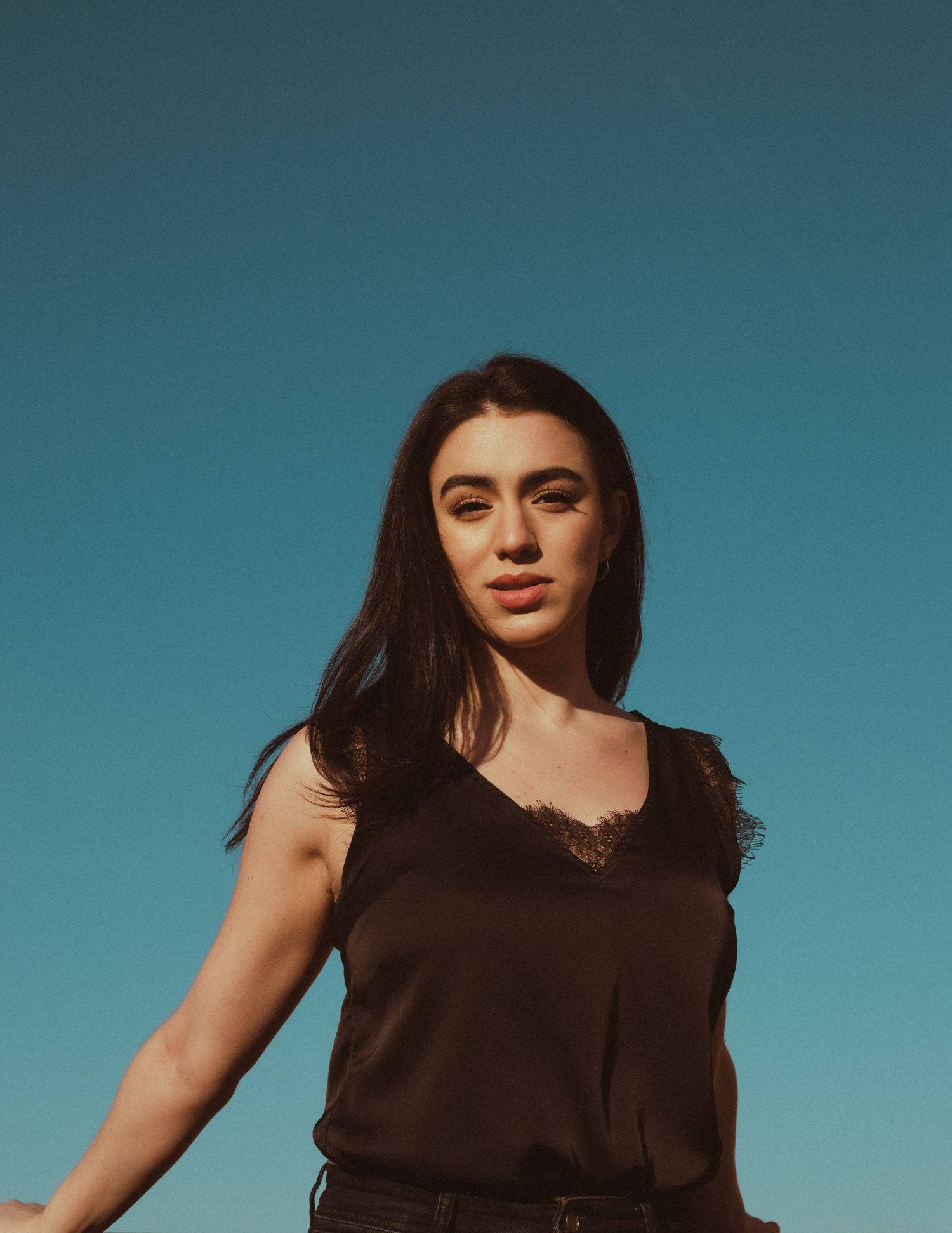 Young woman with long dark hair wearing a black sleeveless top poses against a clear blue sky.