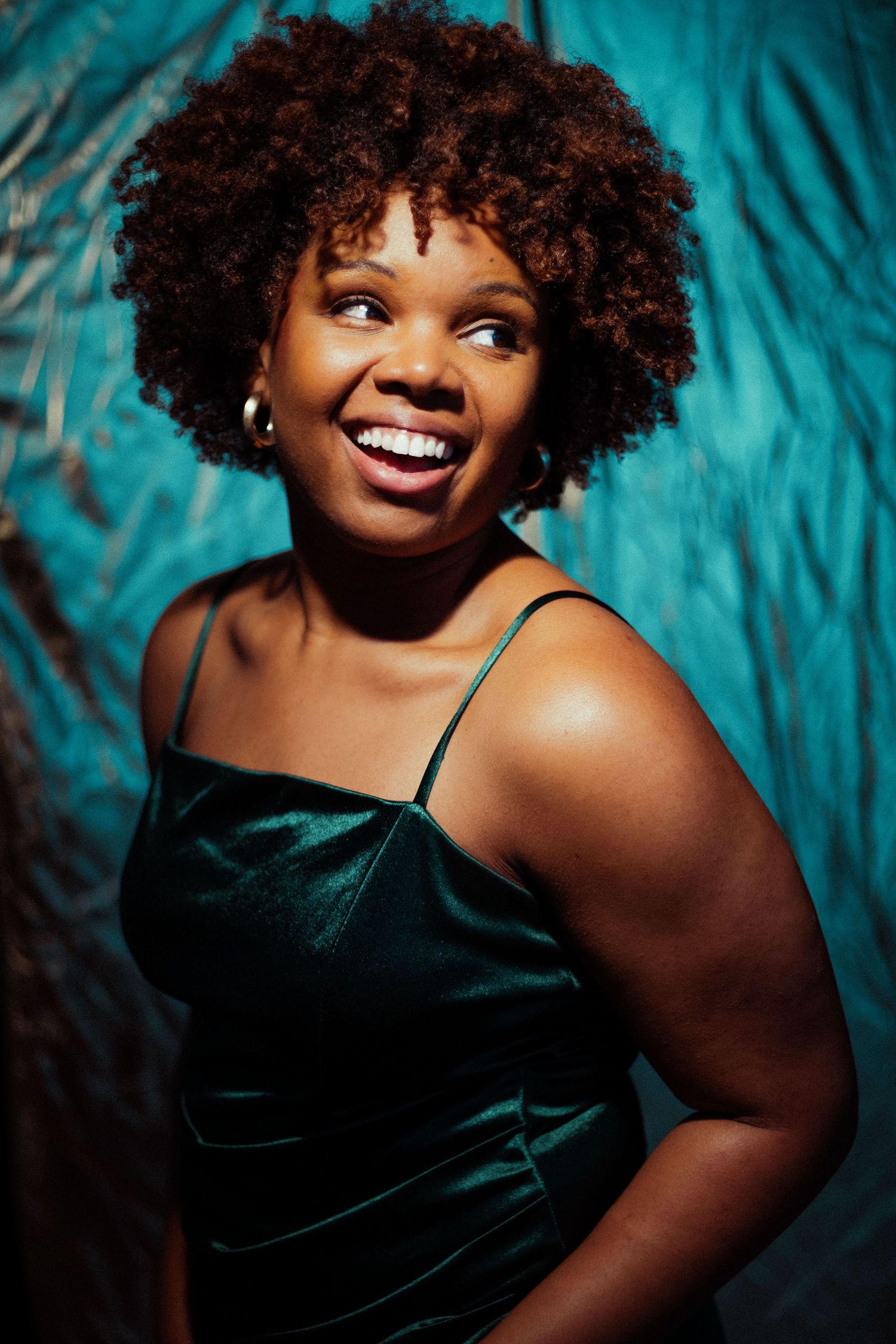 Smiling woman with natural afro hair wearing a green velvet dress, posing against a teal textured background.