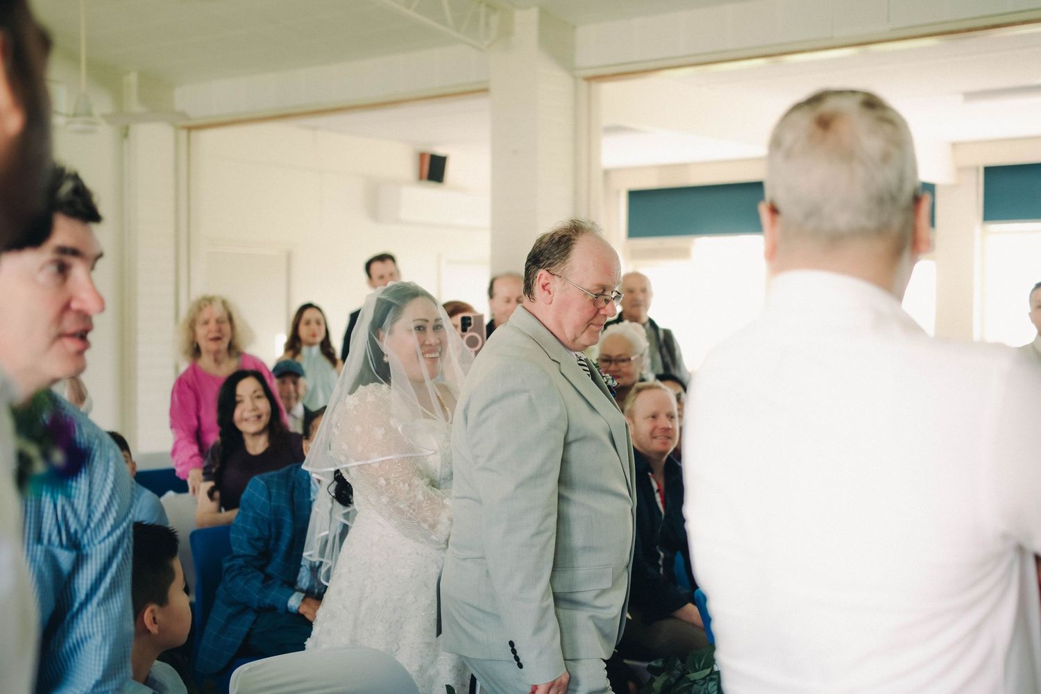 Bride in white veil walks down aisle with escort during indoor wedding ceremony.