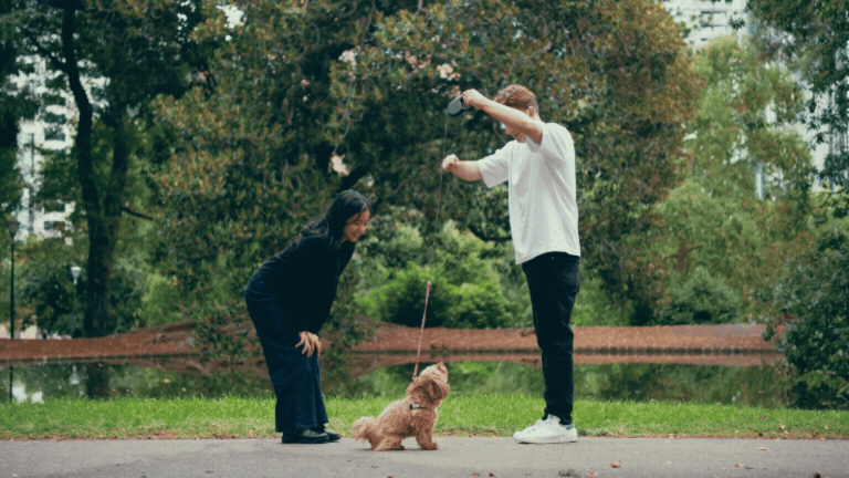 Couple playing with a fluffy dog on a leash in a lush green park near a tranquil pond.