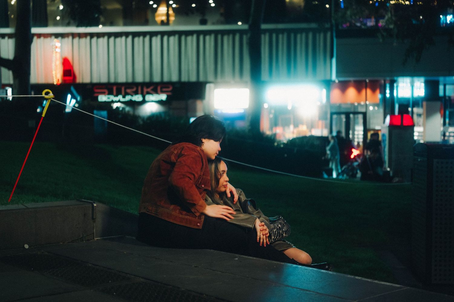Couple sitting closely together on a grassy area outside a bowling alley at night.