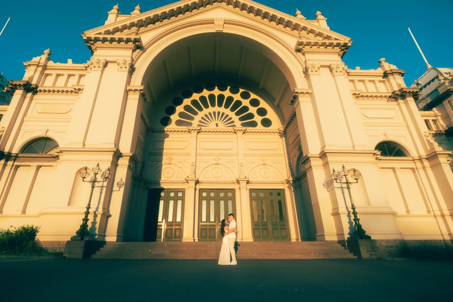 Couple embracing in front of a grand ornate building with large arched entrance at golden hour.