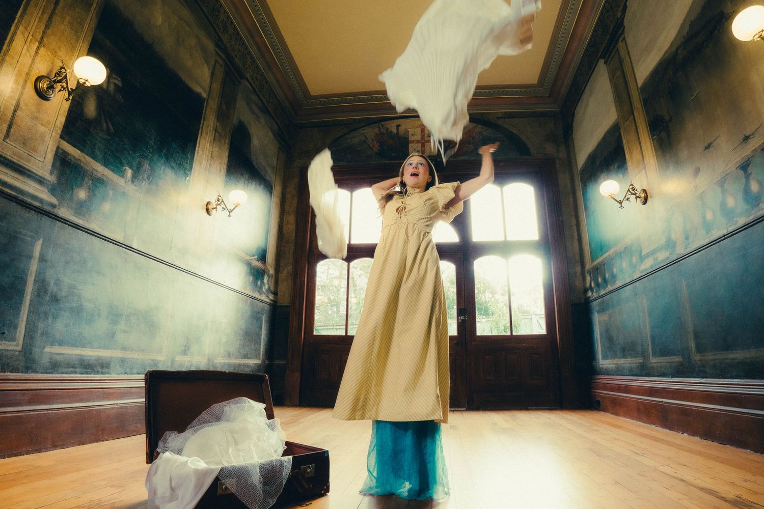 Woman in a vintage dress joyfully tossing fabric in an elegant historic room.