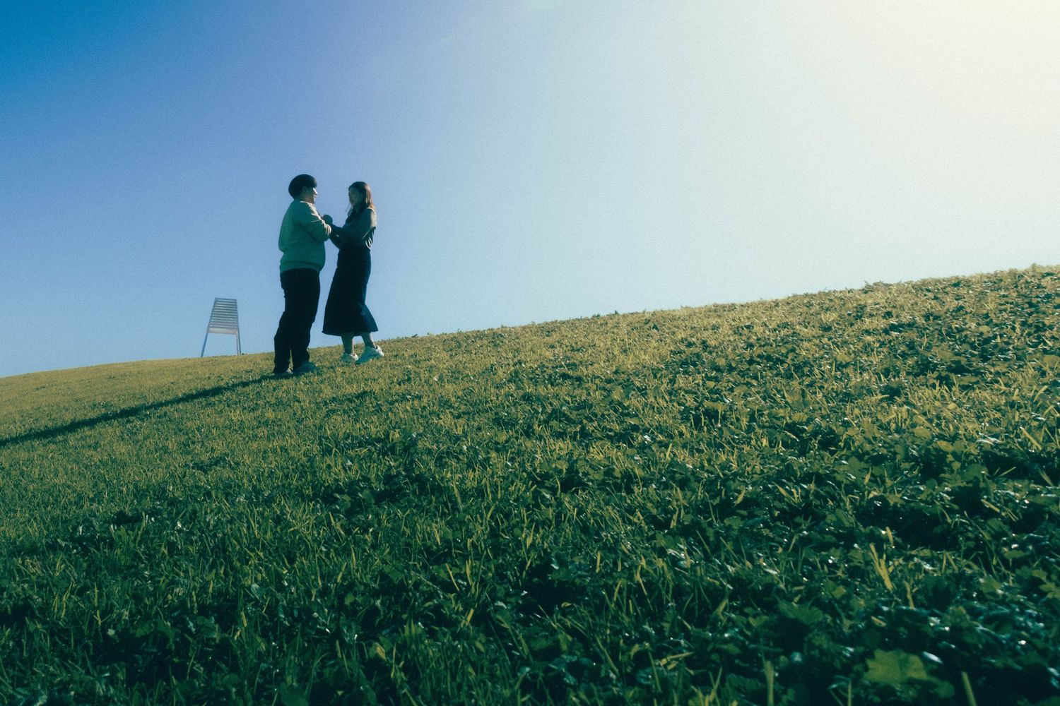 Two people standing on a sunny green hillside, silhouetted against a clear blue sky.