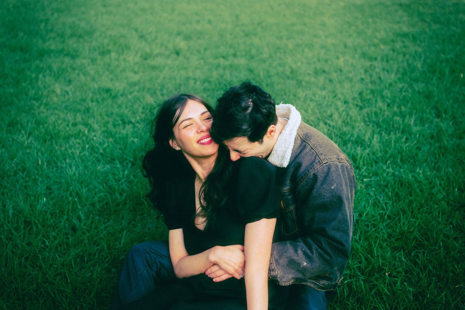 Couple sitting on green grass, woman laughing joyfully as man leans in close, sharing a tender romantic moment outdoors.