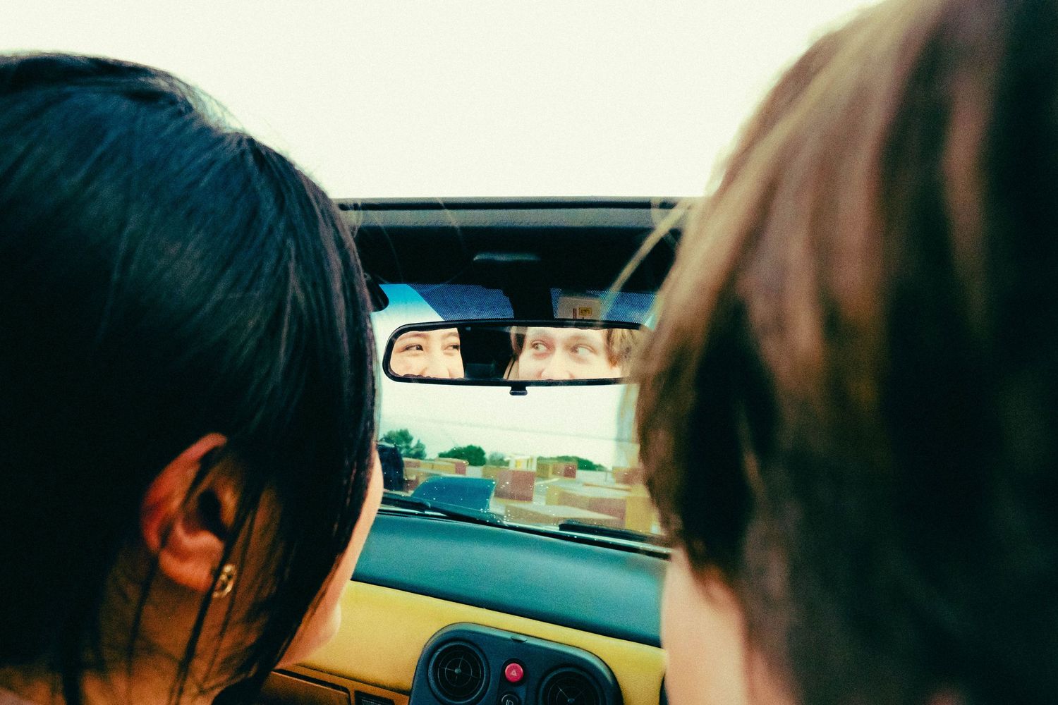 Two passengers view the road ahead from inside a yellow car, with eyes reflected in the rearview mirror.