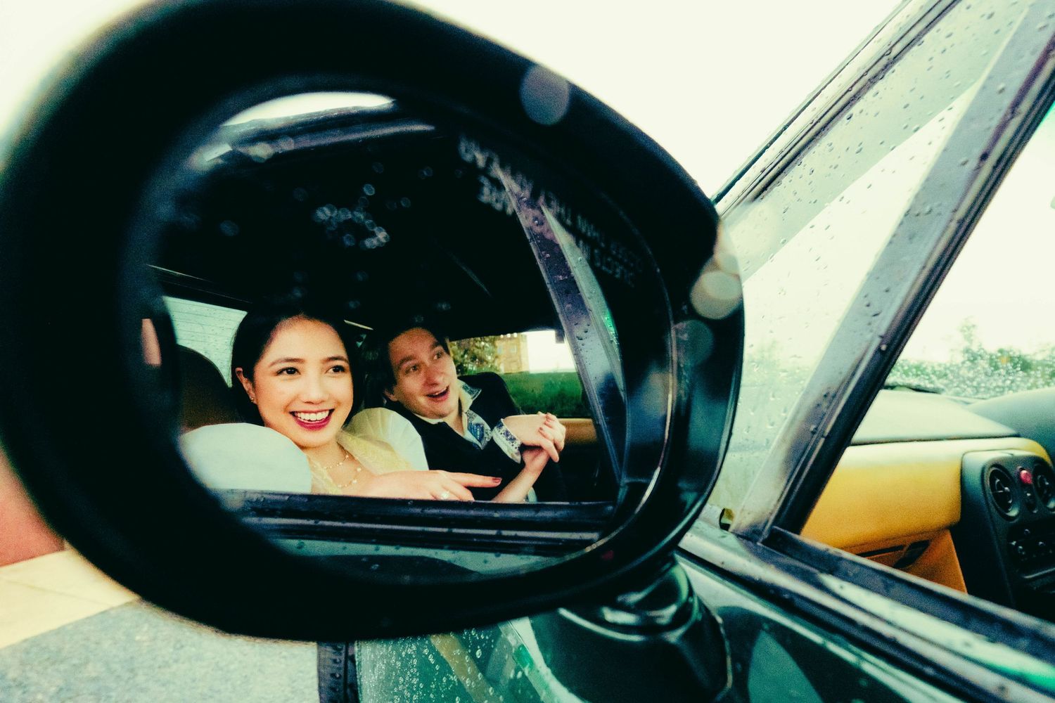 A smiling couple reflected in a car's side mirror on a rainy day.