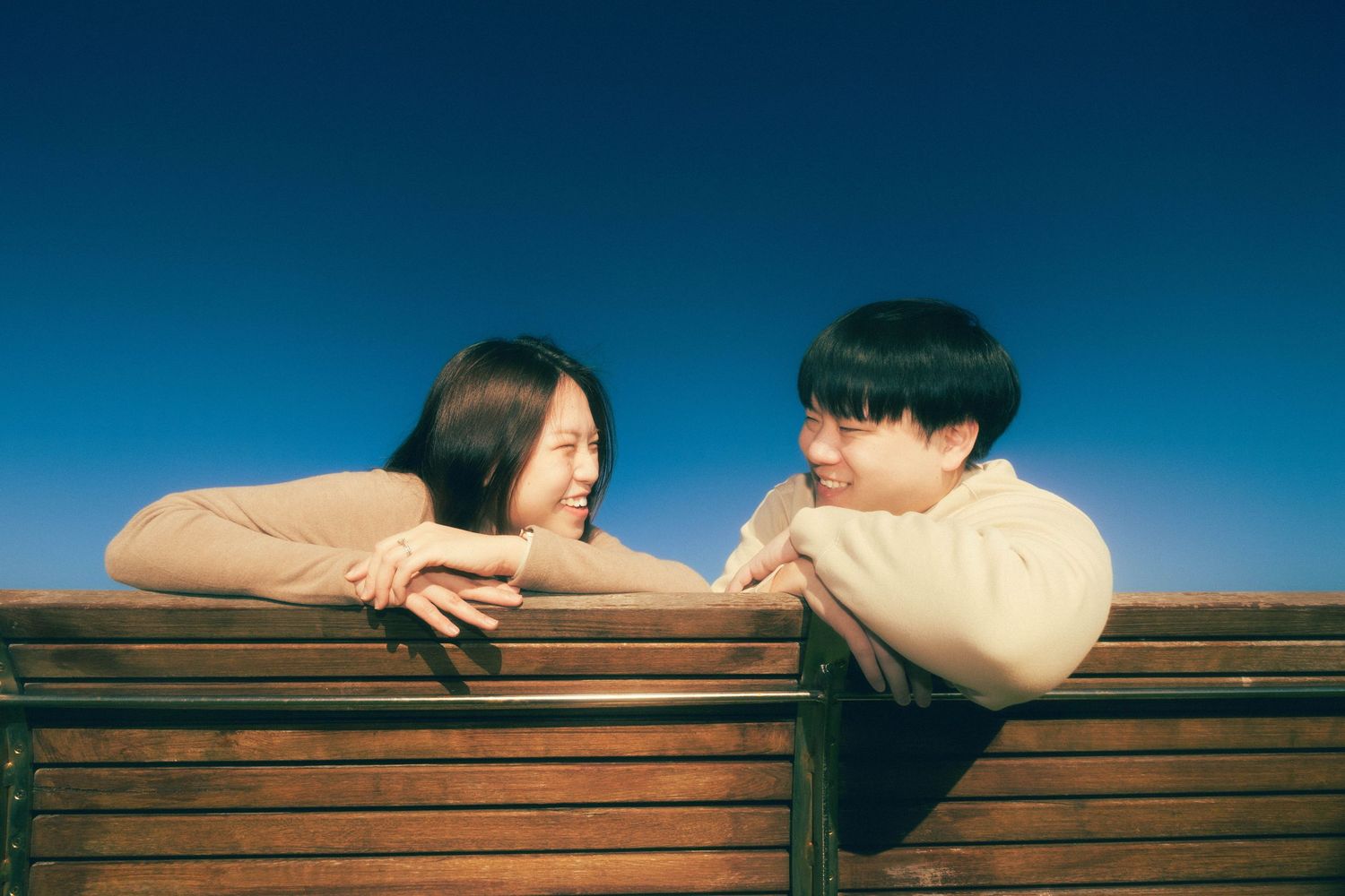 Two young people lean on a wooden bench, smiling at each other against a clear blue sky.