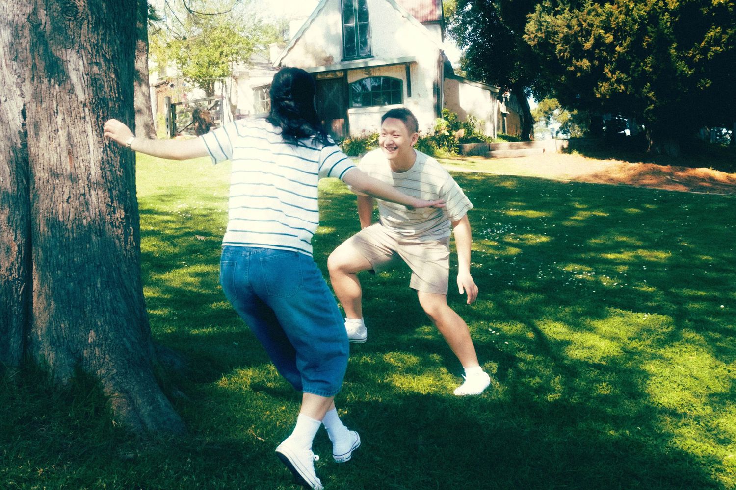 Two people playfully dancing on a sunny green lawn near a large tree in a residential backyard.