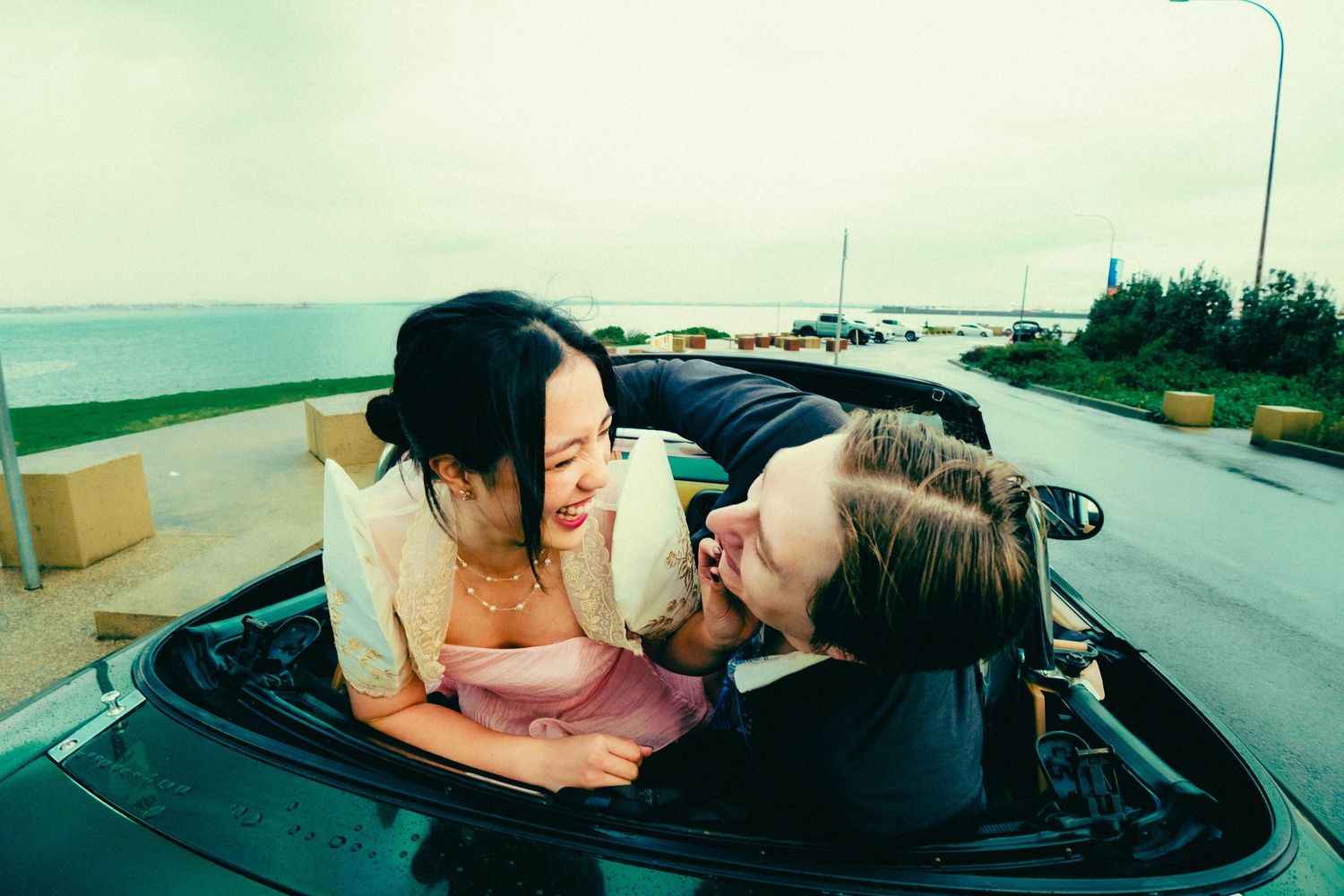 A couple shares a tender kiss in a black convertible parked by the seaside.