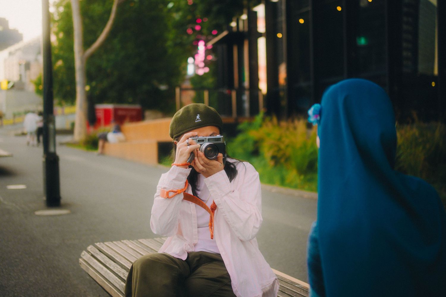 Person sitting on bench photographing woman in blue hijab on urban tree-lined path.