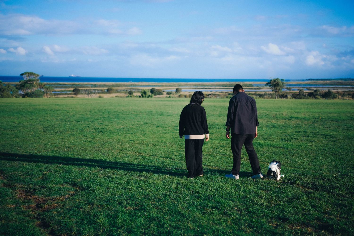Two people in black outfits walking a small dog on a lush green field by the ocean.