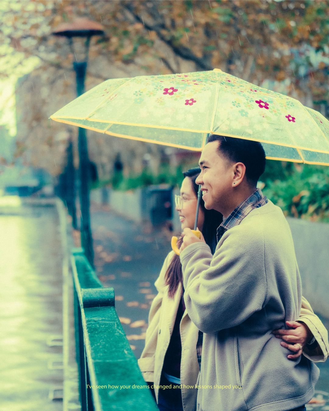 Couple sharing an umbrella in the rain during autumn, embracing by a waterway lined with colorful foliage.