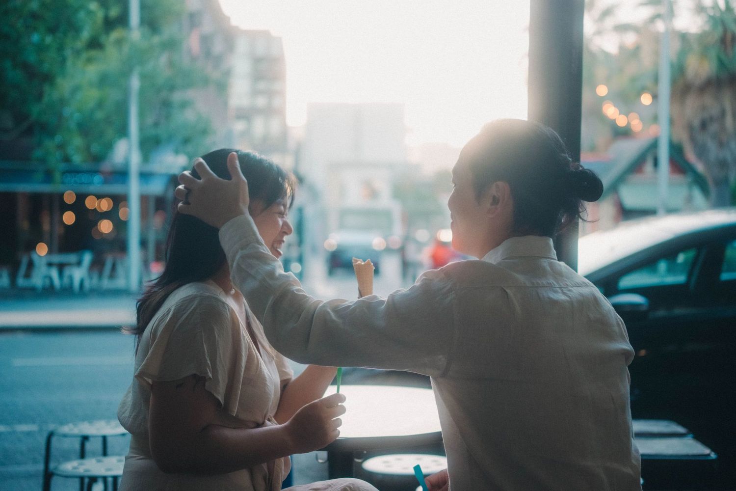 Man gently fixing a woman's hair at an outdoor cafe table on a busy city street at dusk.