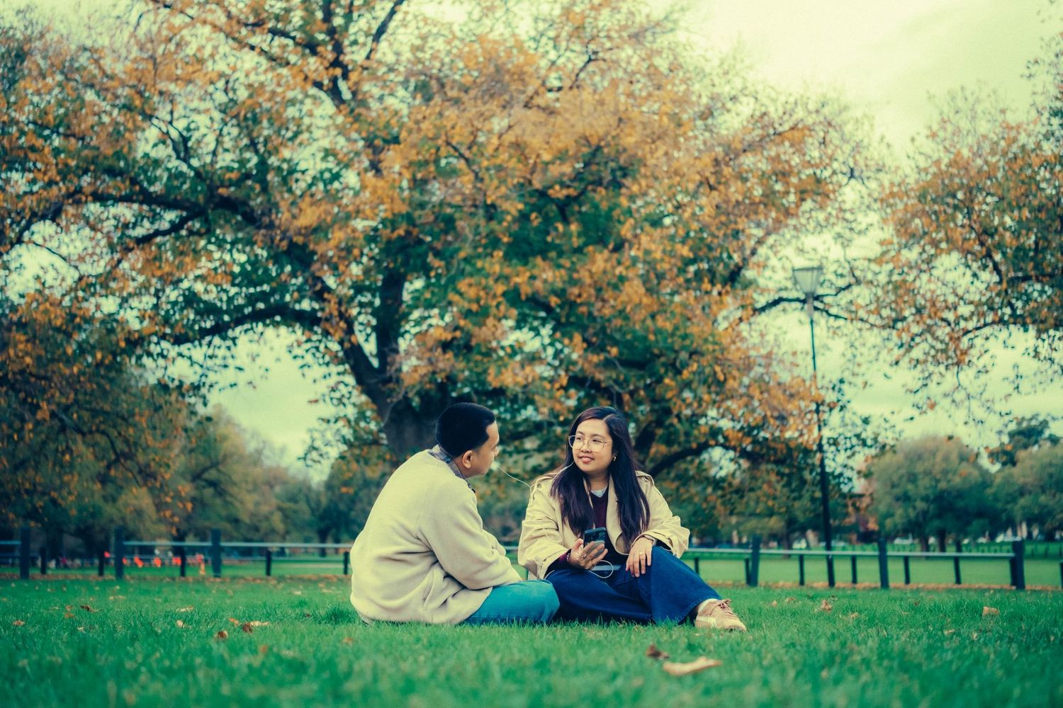 Couple sitting on grass in a park having a conversation beneath a colorful autumn tree.