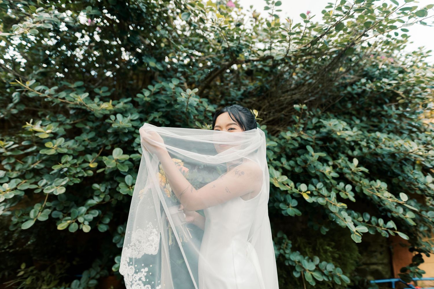 A bride holds up her lace wedding veil in front of lush green trees outdoors.