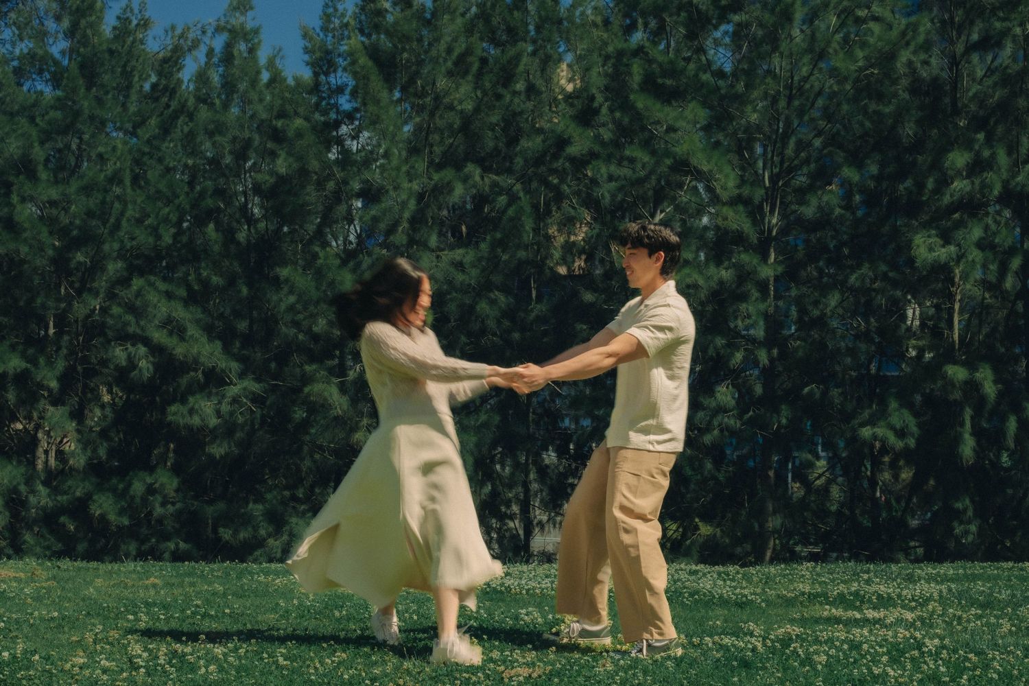Happy couple holding hands and spinning in green meadow surrounded by pine trees.